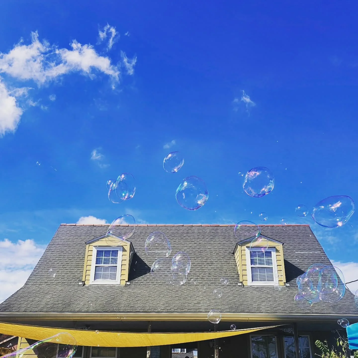 Bubbles floating in the sky above a house with two dormer windows, a shingled roof, and a yellow awning, with a blue sky and some clouds in the background.