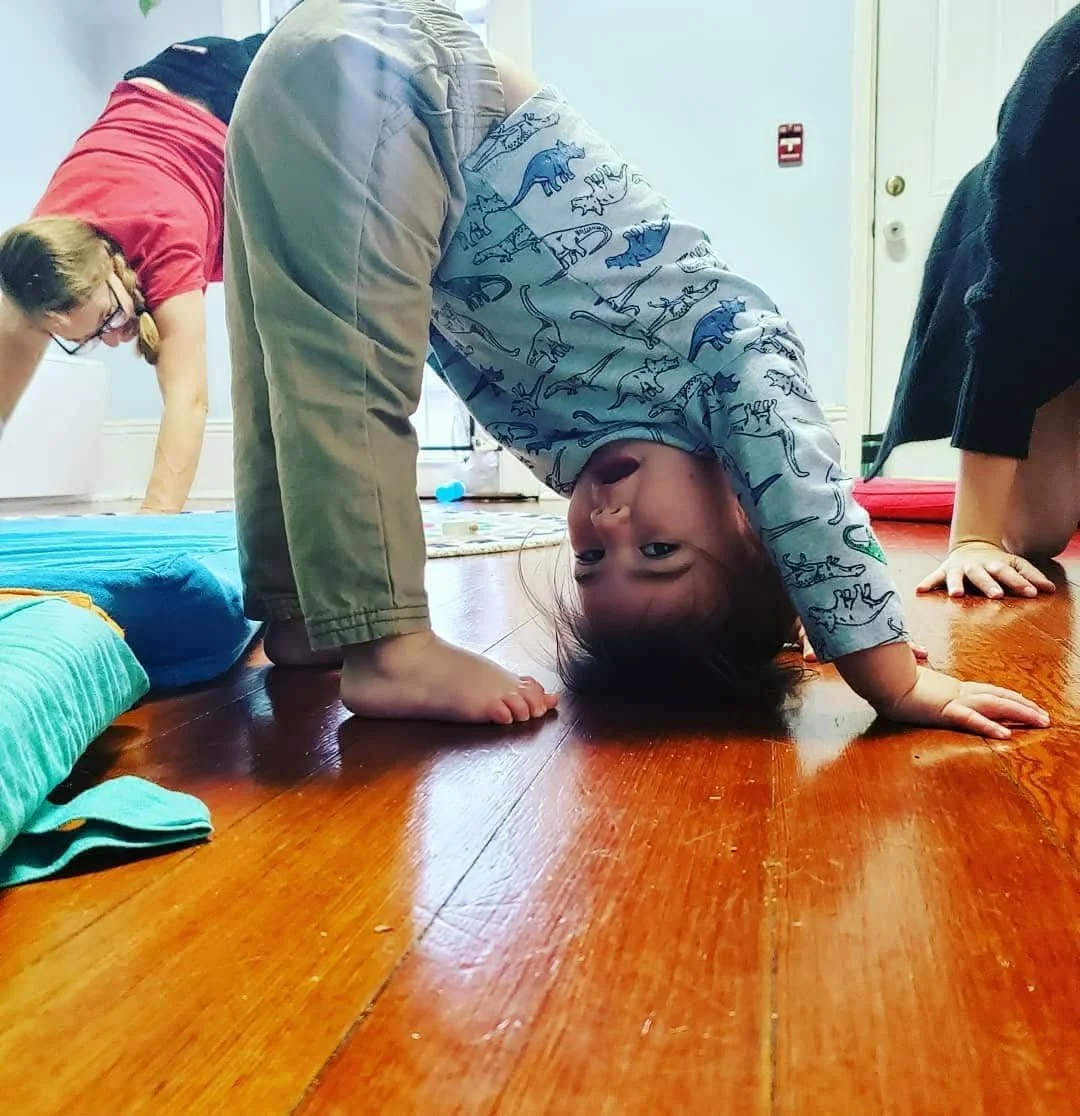 A young boy in a dinosaur print shirt is upside down in a bridge pose on a wooden floor, smiling at the camera. Two other children are partially visible nearby, one in a red shirt and another in black, in a room with white walls and a door in the background.