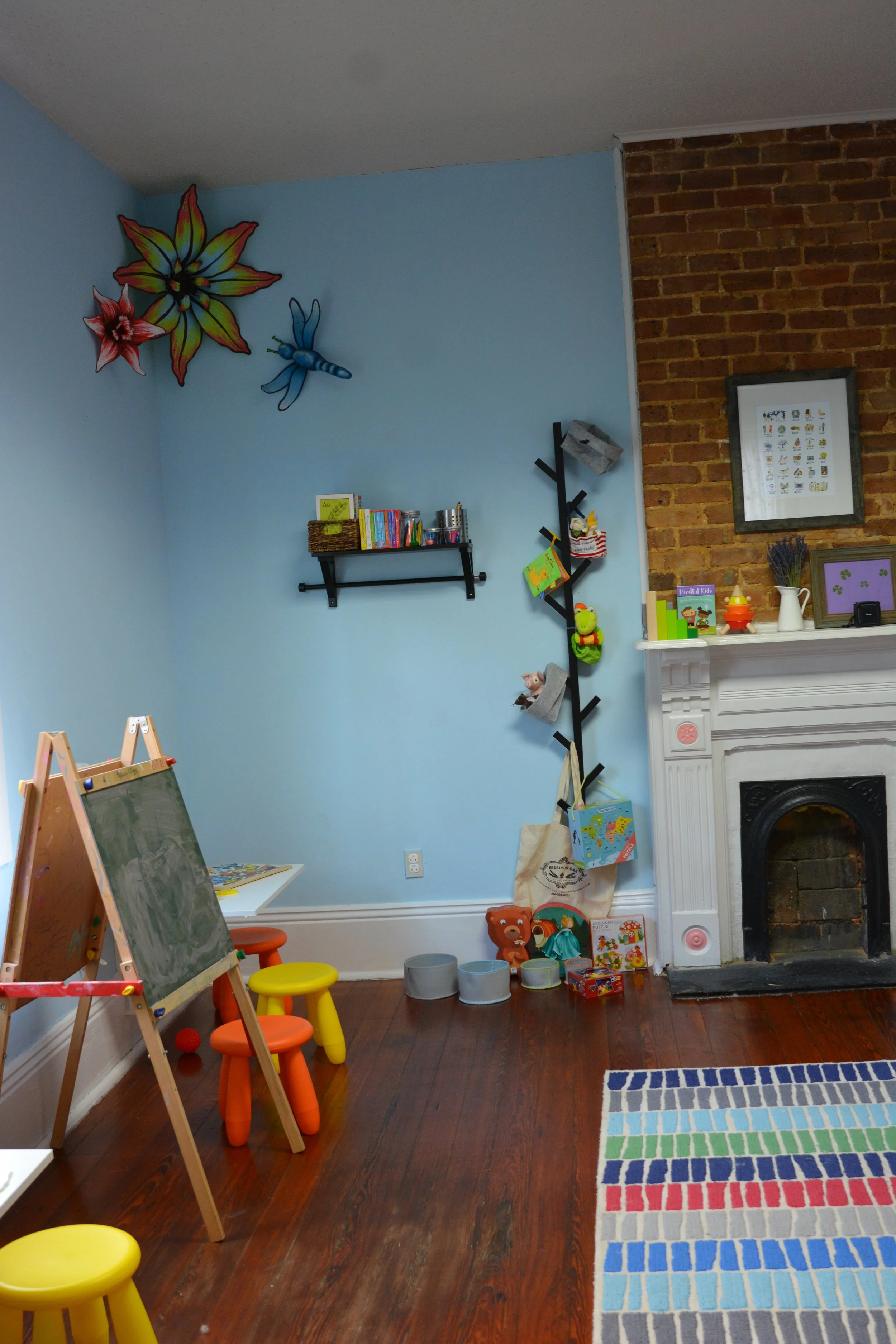 A children's playroom with blue walls, decorated with large colorful flower and insect wall decals. There is a small chalkboard easel, bright yellow and orange stools, a rainbow color brick-patterned rug, a white fireplace with children's books, toys