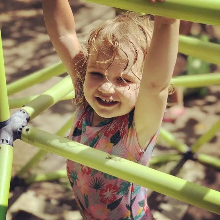 A young girl with curly blonde hair and a flower-patterned shirt hangs from a yellow jungle gym outdoors, smiling at the camera.