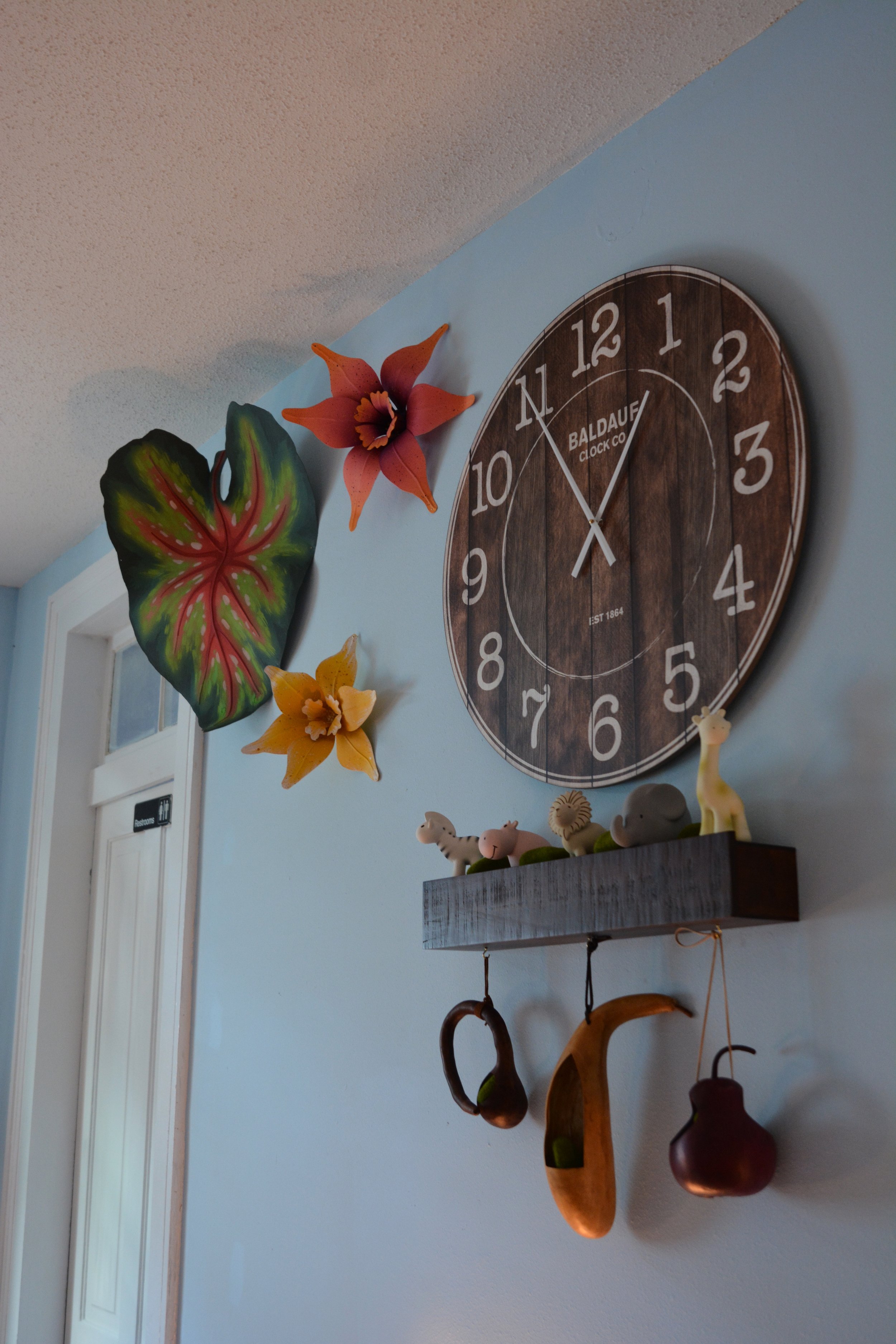 Decorative items on a light blue wall including a wooden clock with white numbers and a black second hand, artificial flowers resembling a lily, a patterned leaf, and small animal figurines on a gray wooden shelf with hanging brown and wooden hooks.