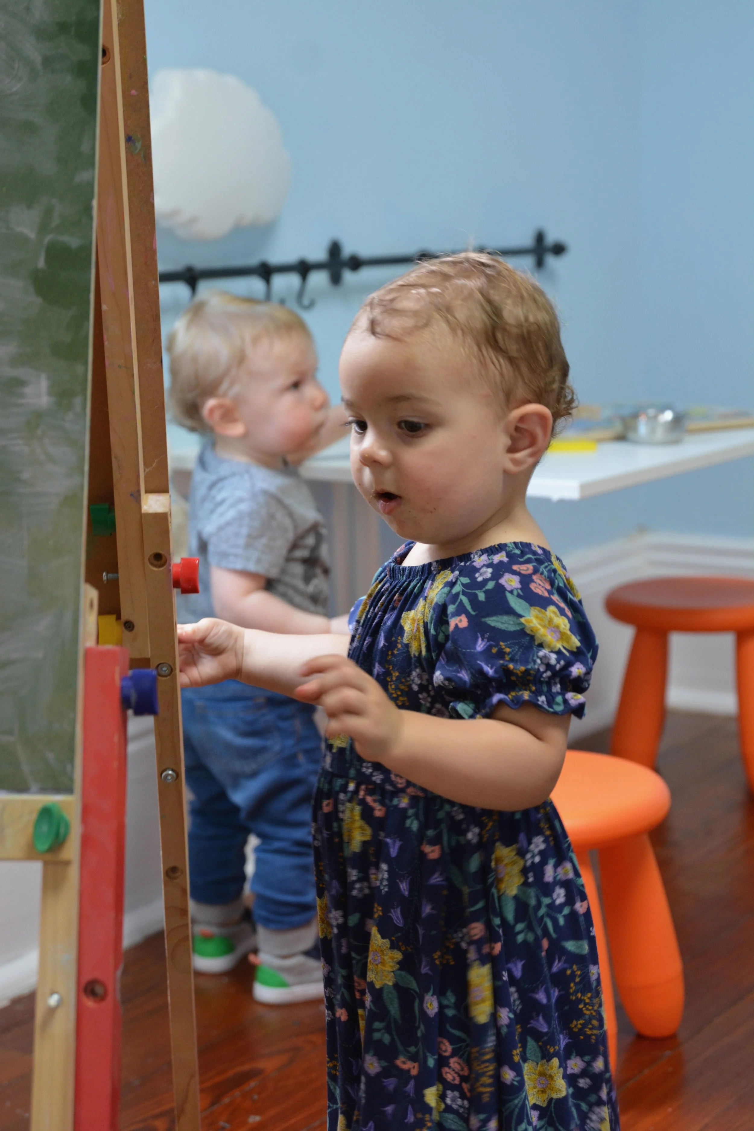 Two young children, a girl with a floral dress and a boy with a gray shirt, are playing at an art easel. They are focused on their activity in a room with a blue wall and wooden floor.