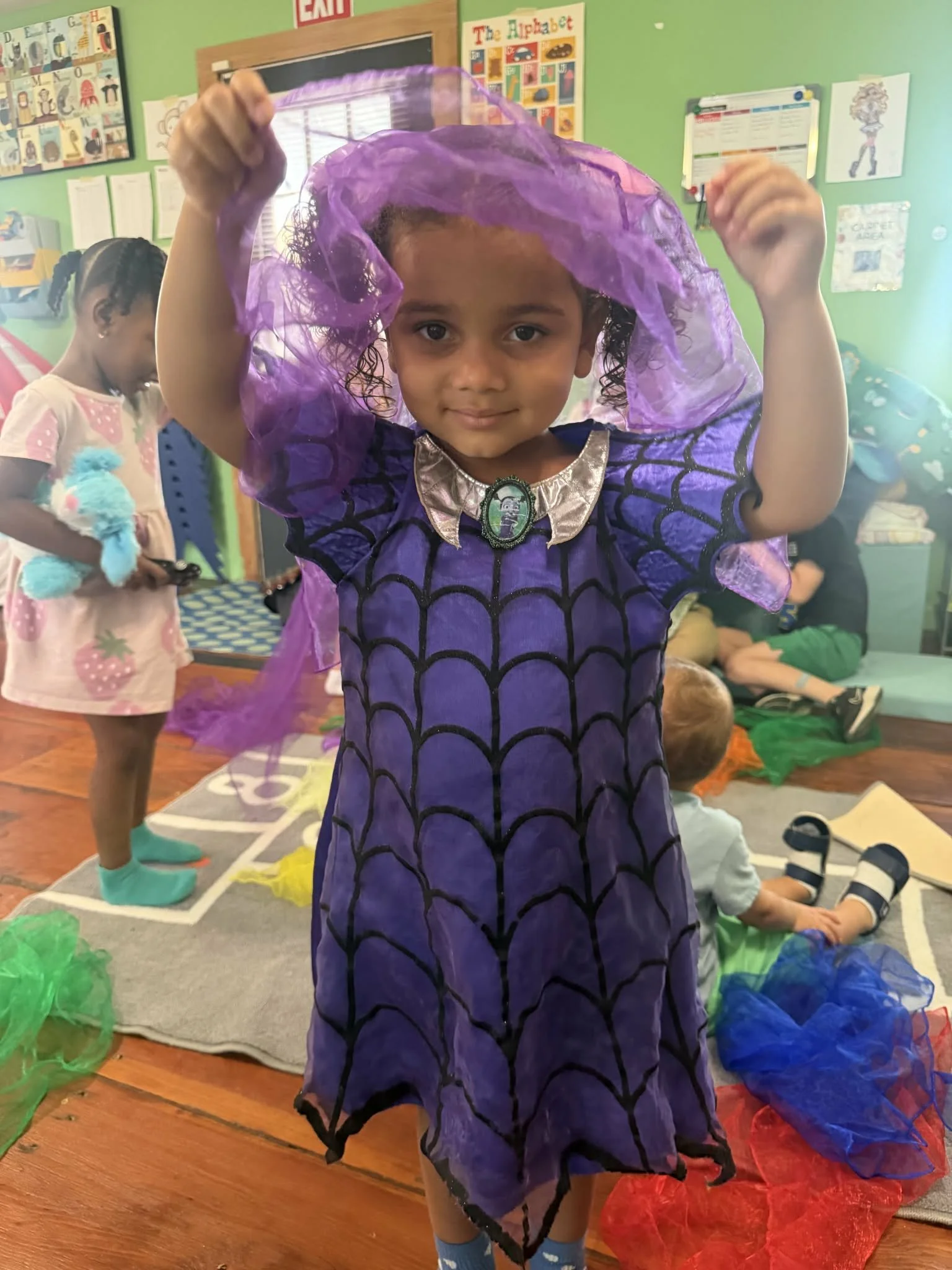 A young girl in a purple costume with a web pattern is holding purple tissue paper above her head. Other children are in the background with colorful tissue paper on the floor in a classroom setting.
