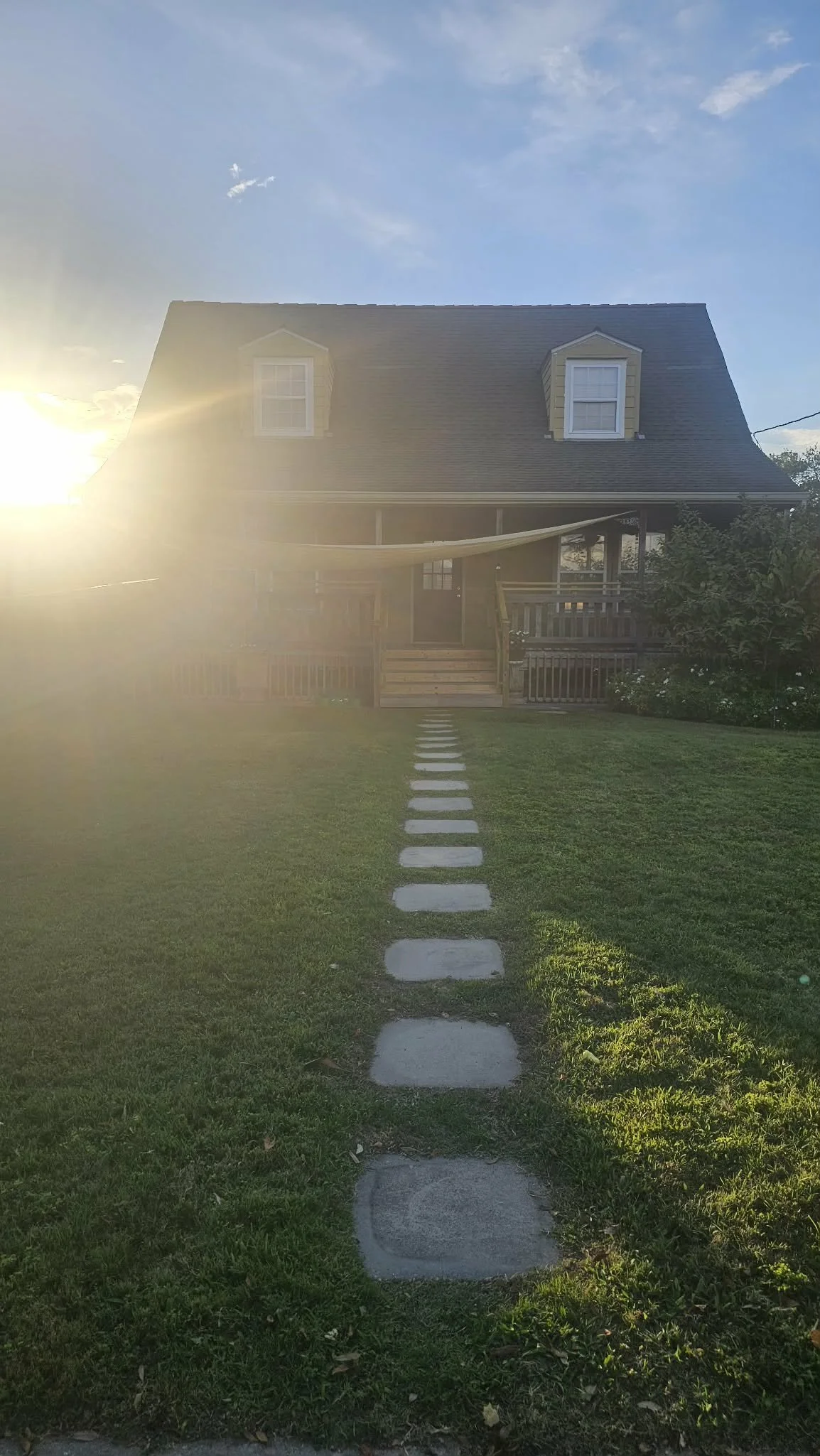Sunset view of a house with two dormer windows in the roof, a porch, and a stone pathway leading to the front door, with a grassy lawn and bushes on the sides.