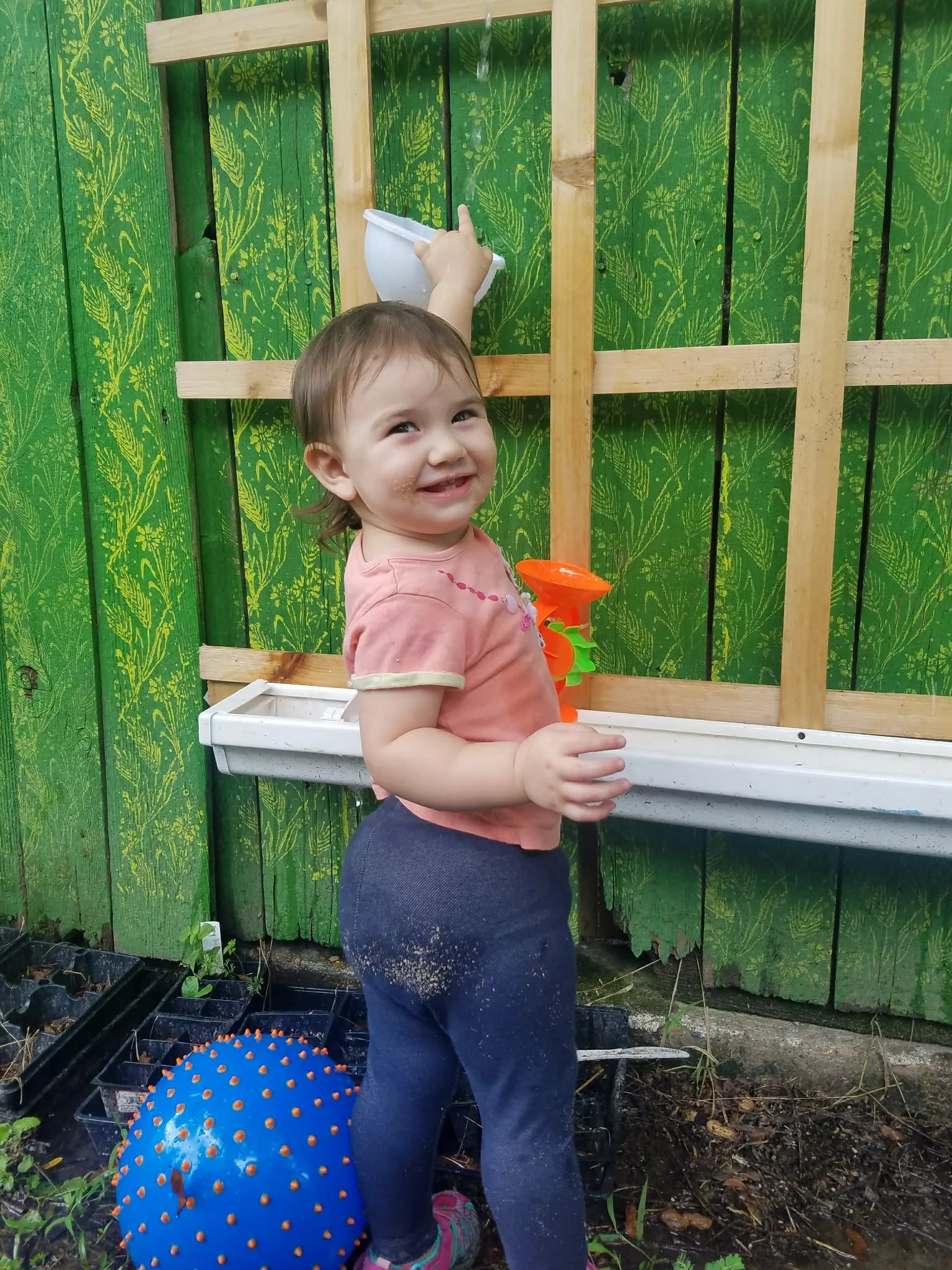 A young girl with wet hair smiling and pointing at a bird bath on a green wooden fence with a yellow pattern. She is wearing a pink shirt and dark pants, and there is a blue textured ball next to her on the ground. A container with potted plants and some gardening tools are also visible.