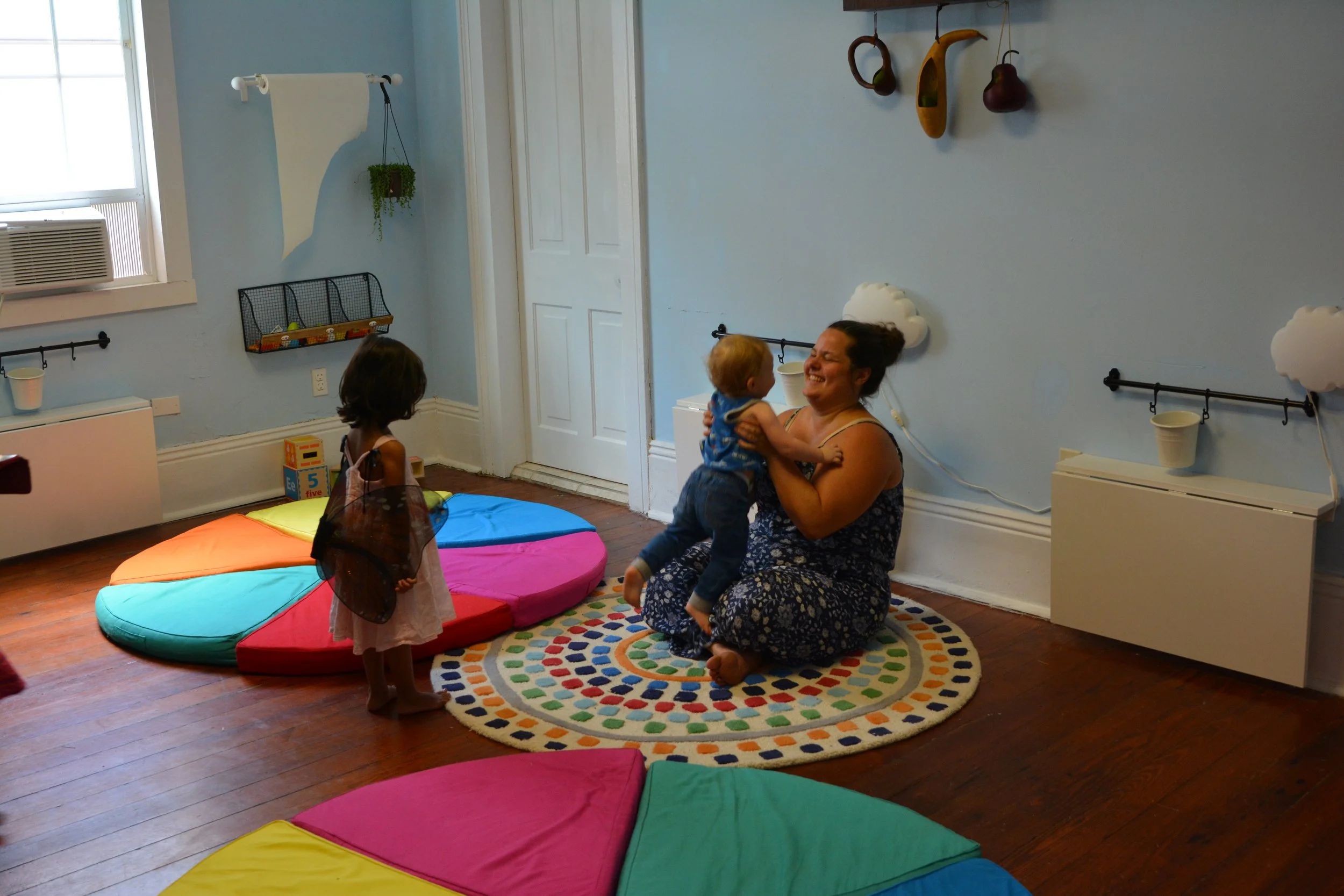 A woman smiling and kneeling on a colorful rug while holding a young child, with a young girl standing nearby in a playroom with rainbow-colored foam mats and wall decorations.