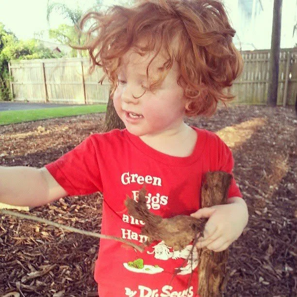 A young child with curly red hair playing outside, holding a small bird and a wooden stick, standing in a yard with a fence and trees in the background.