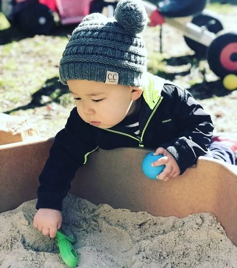 Young child in a knit hat playing in a sandbox, holding a green shovel and a blue ball, with a toy car and a stroller in the background.