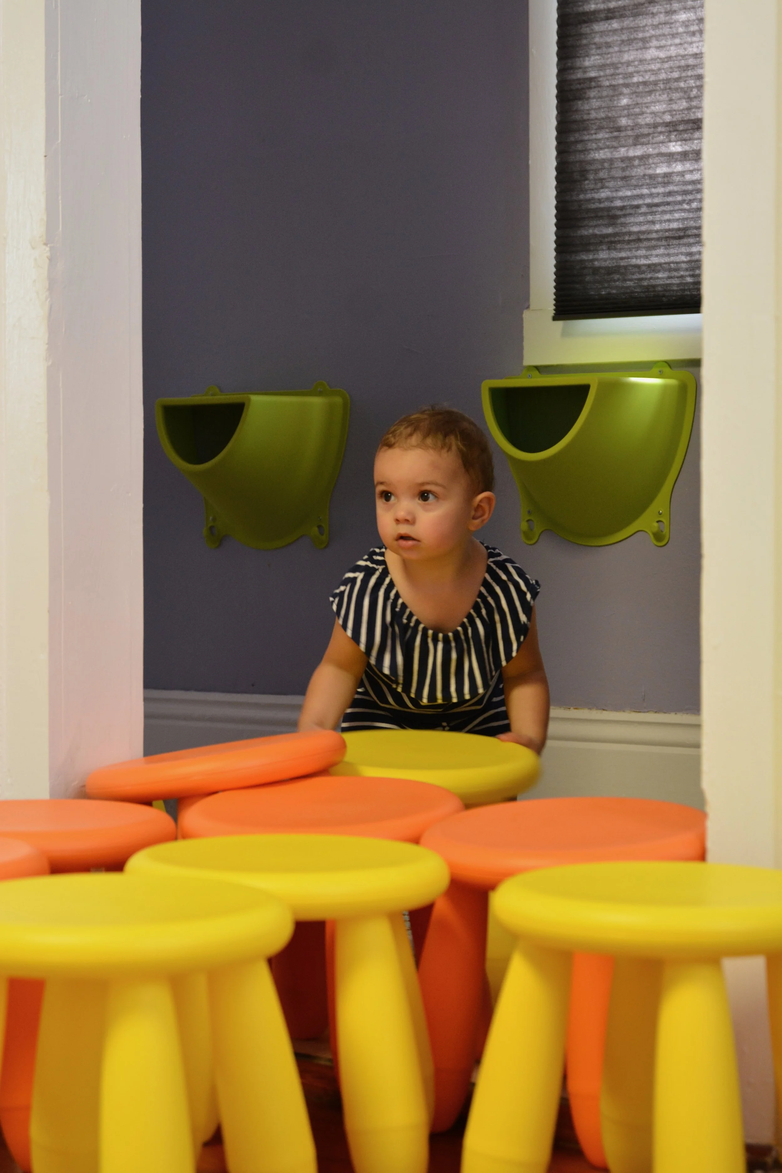 A young child with short brown hair, wearing a black and white striped shirt, is seen among multiple small orange and yellow circular stools arranged in a playful setting.
