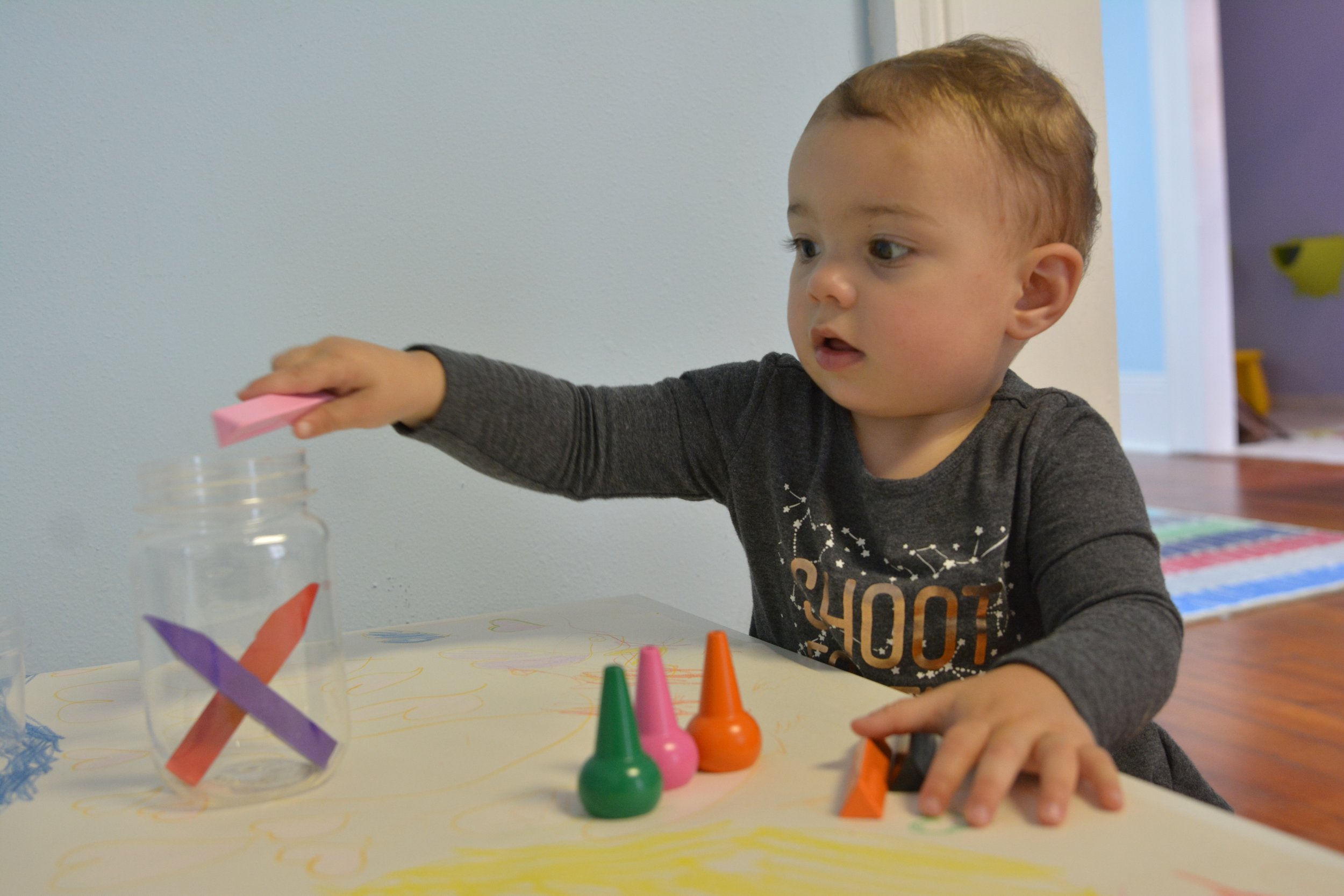A young child with short brown hair, wearing a gray shirt with white text and designs, is reaching out with his right hand towards colorful objects on a table. The table has various craft materials, including small cones, paper, and a transparent cup