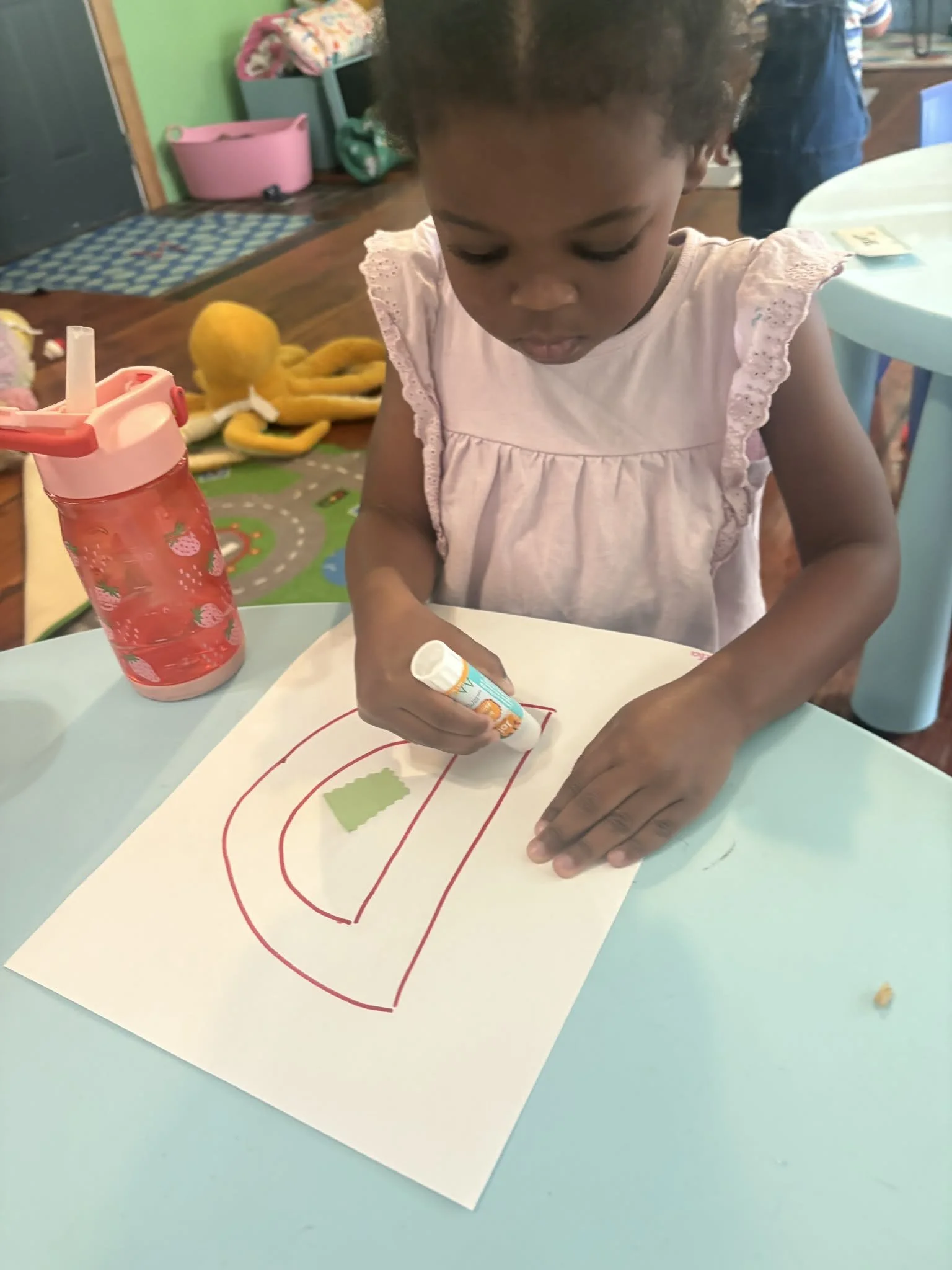 A young girl with dark curly hair is coloring on a sheet of paper with red and green lines, using a glue stick. She is wearing a pink shirt with lace details on the sleeves. There is a pink water bottle with a straw on the table, and a yellow plush t