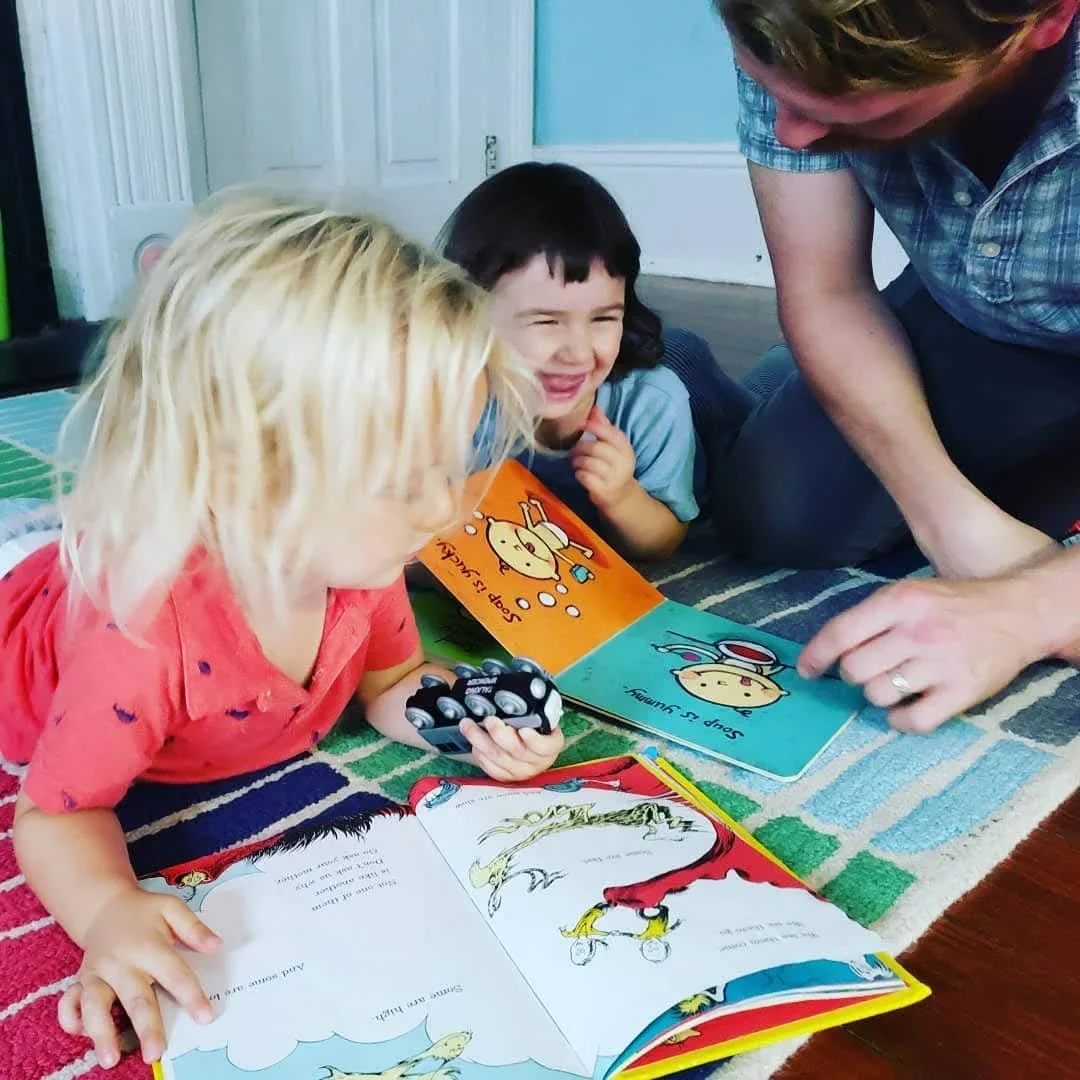 Two children and an adult reading books and looking at toys on a colorful rug in a room with a door and light blue walls.