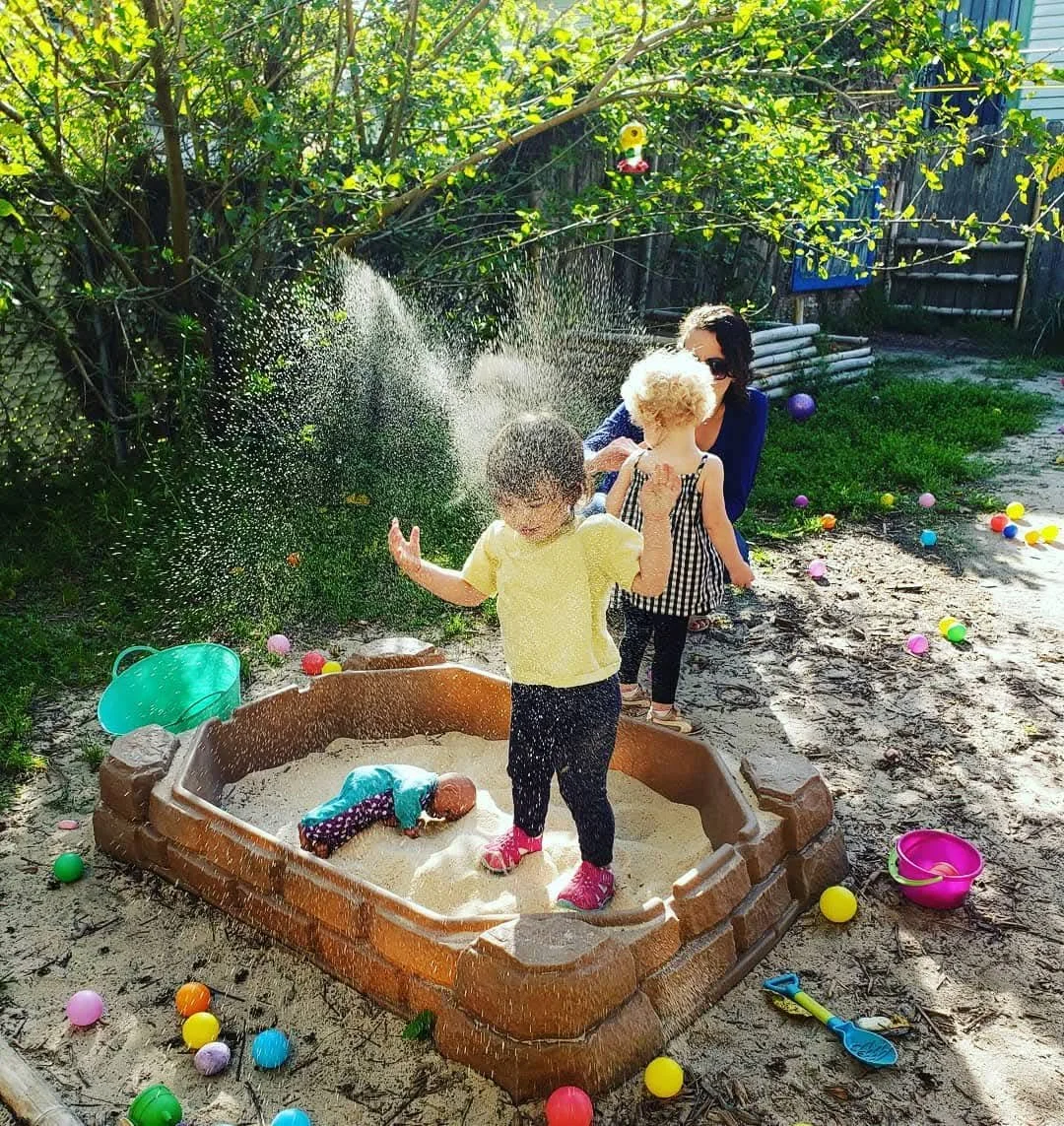 Children playing in a sandbox with colorful plastic eggs, while their mother watches and helps, surrounded by toys and a backyard with trees.