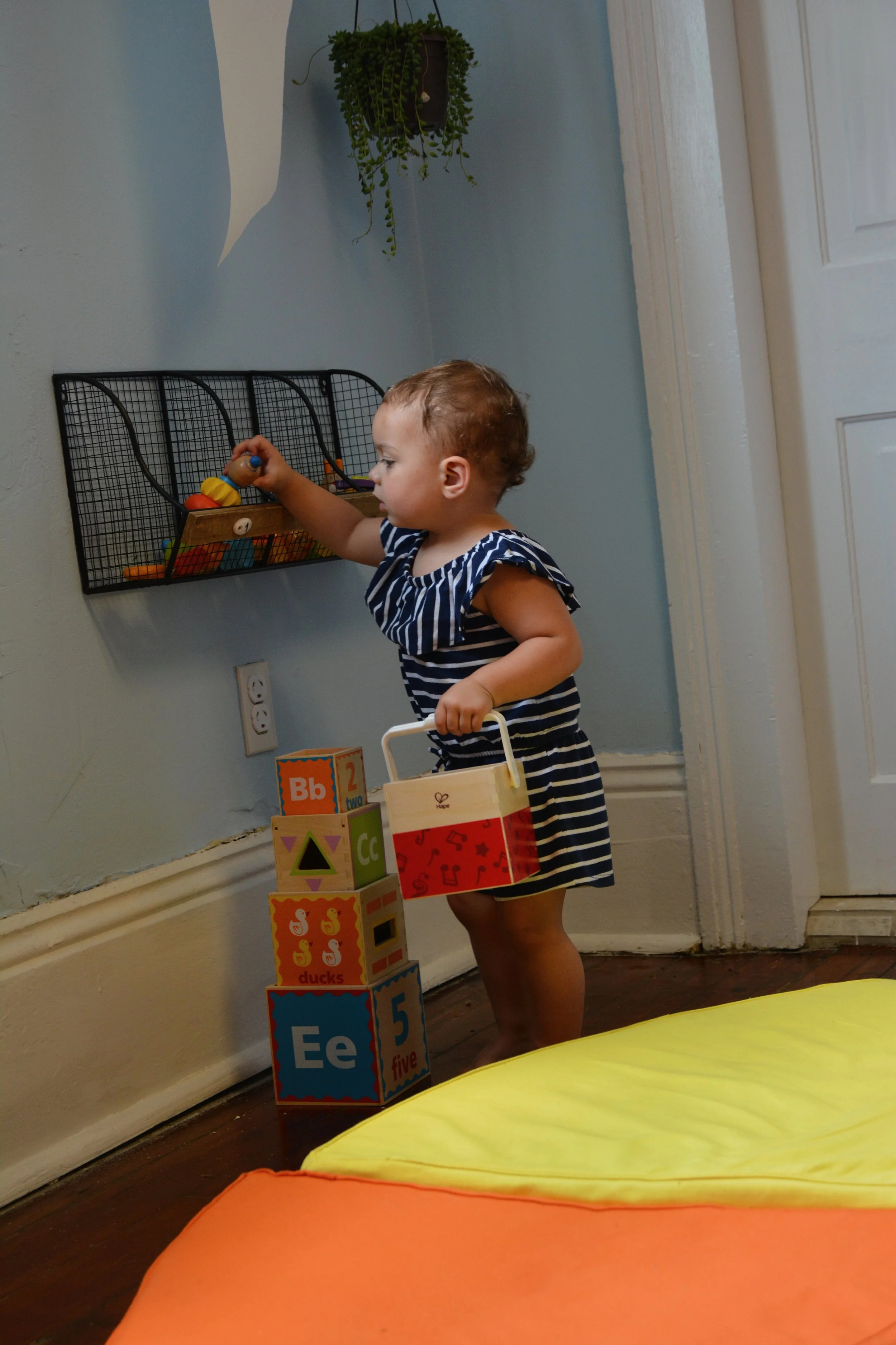 A young child wearing a navy blue and white striped dress stacking colorful educational blocks with letters and numbers on a hardwood floor in a room with light blue walls and a white door. There is a yellow and orange cushion in the foreground and a
