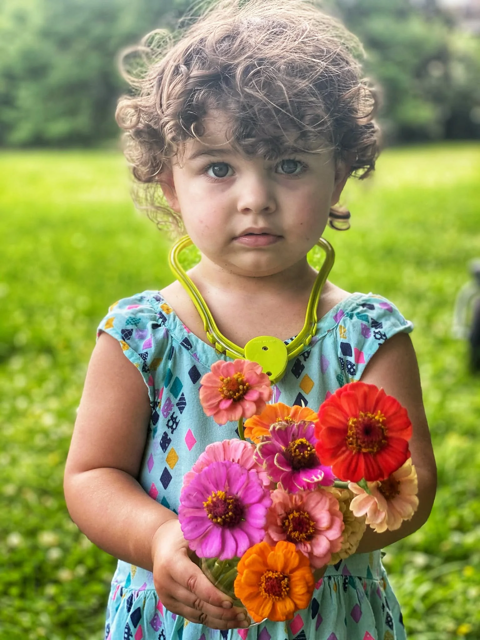 A young girl with curly hair, wearing a colorful dress, holds a bouquet of bright pink, orange, and red flowers, standing outdoors on a green grassy field with trees in the background.