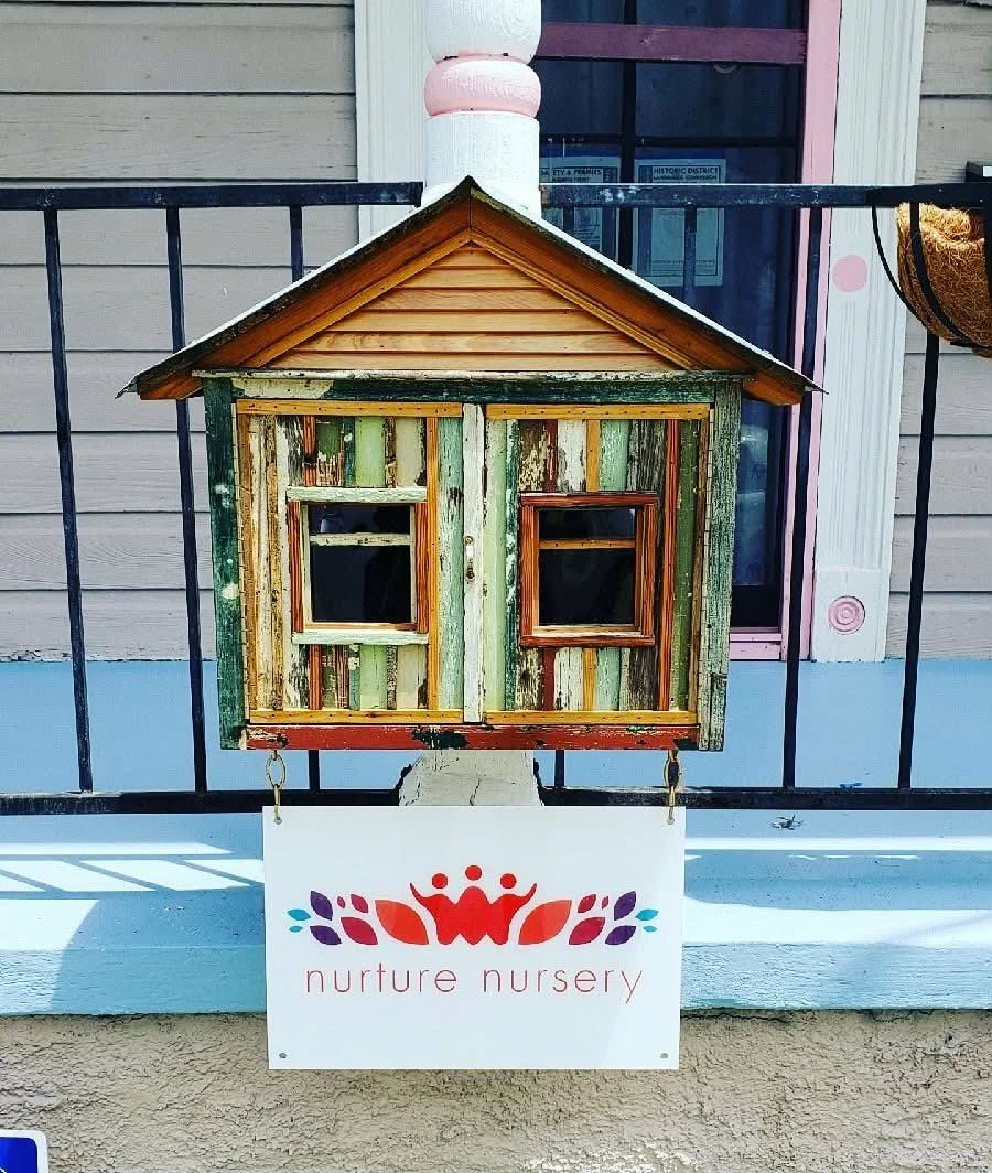 A small decorative wooden house on a porch with a sign underneath that reads 'nurture nursery' featuring a graphic of three red flowers and a red human figure in the center.