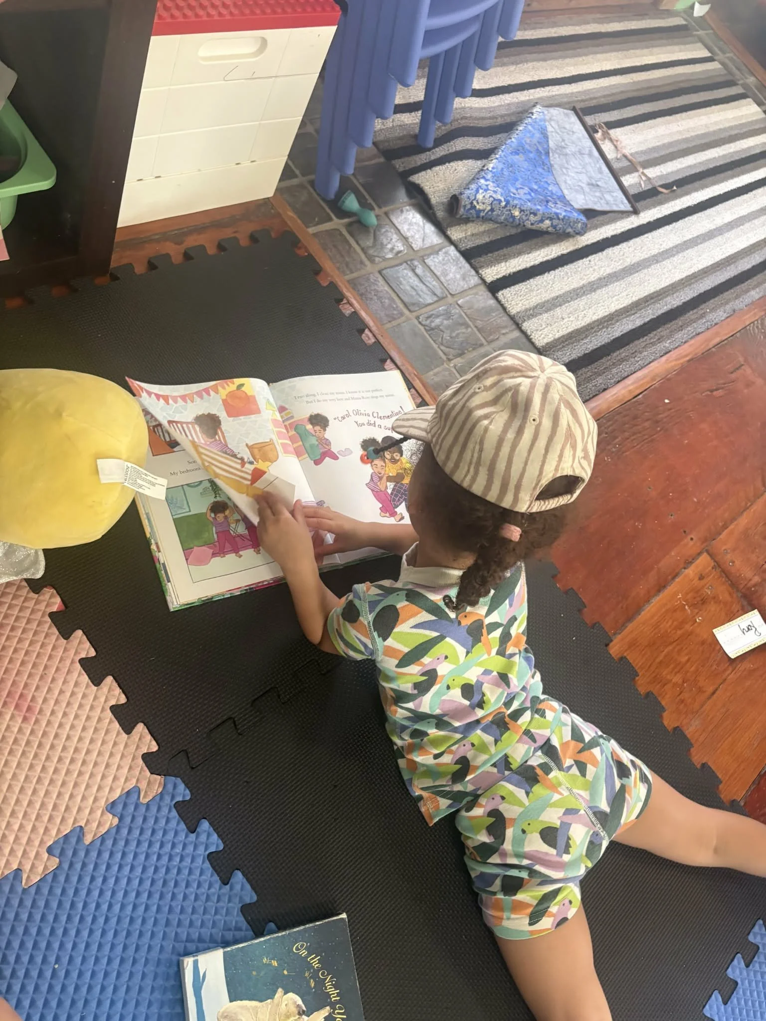 A child wearing a striped pointed cap and colorful pajamas is lying face down on a black foam mat, reading a colorful children's book about a sheep on a striped rug in a room with a tiled floor, a blue table with blue chairs, and a yellow plush toy.