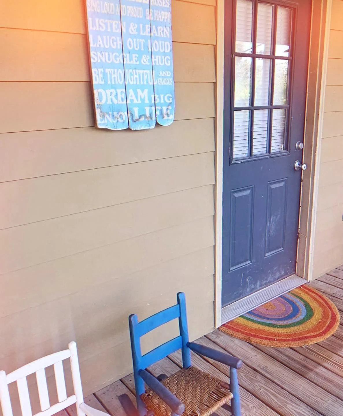 Exterior door of a house with a decorative sign on the wall beside it and a colorful oval rug in front. Two small chairs, one white and one blue with a wicker seat, are on the porch.