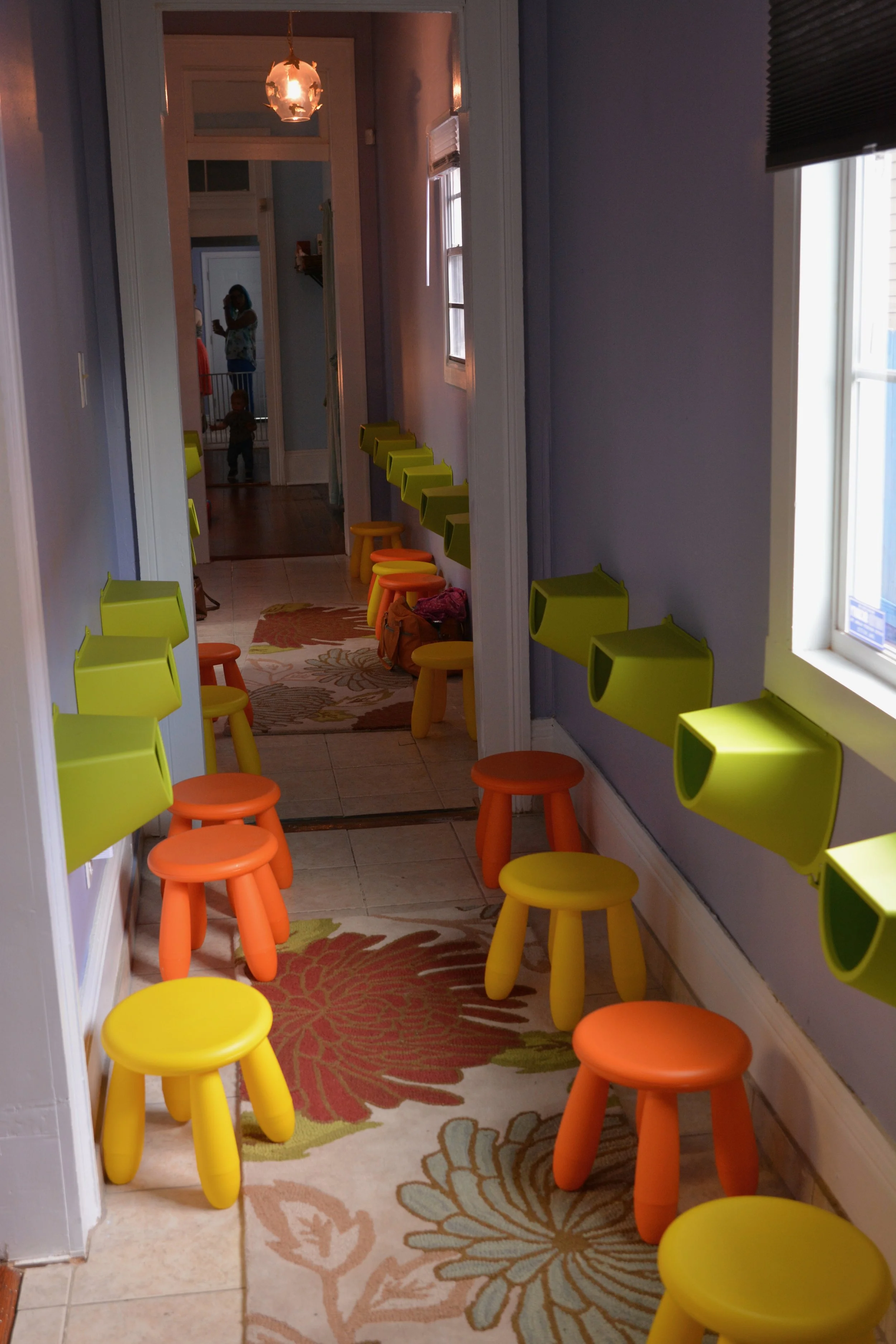 A narrow hallway with a patterned rug, yellow and orange stools, and lime green wall-mounted shelves. The hallway leads to a room with a woman and a child visible in the doorway, and natural light coming through the window on the right.
