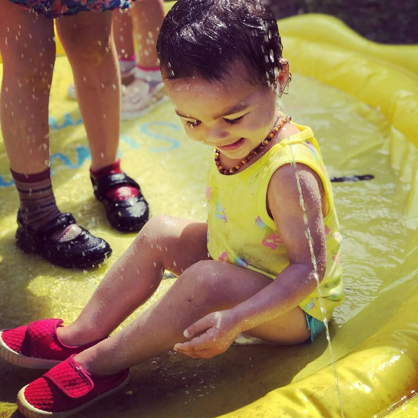 A young girl sitting on a yellow slip-and-slide, smiling, with water and soap on her arms and face, wearing a yellow tank top, shorts, and red shoes, while an adult stands nearby in black sandals and socks.