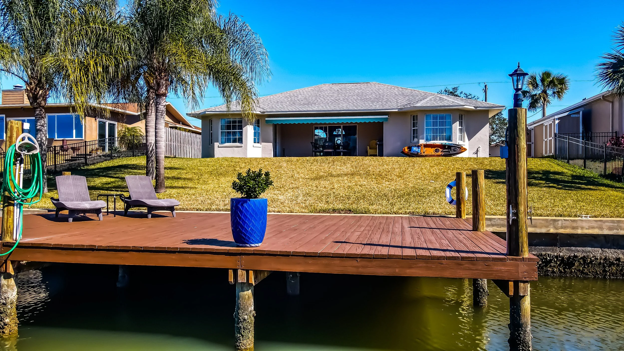 Backyard with a wooden dock leading to a house by the water, two lounge chairs, a blue planter with a shrub, palm trees, and a kayak on the lawn.