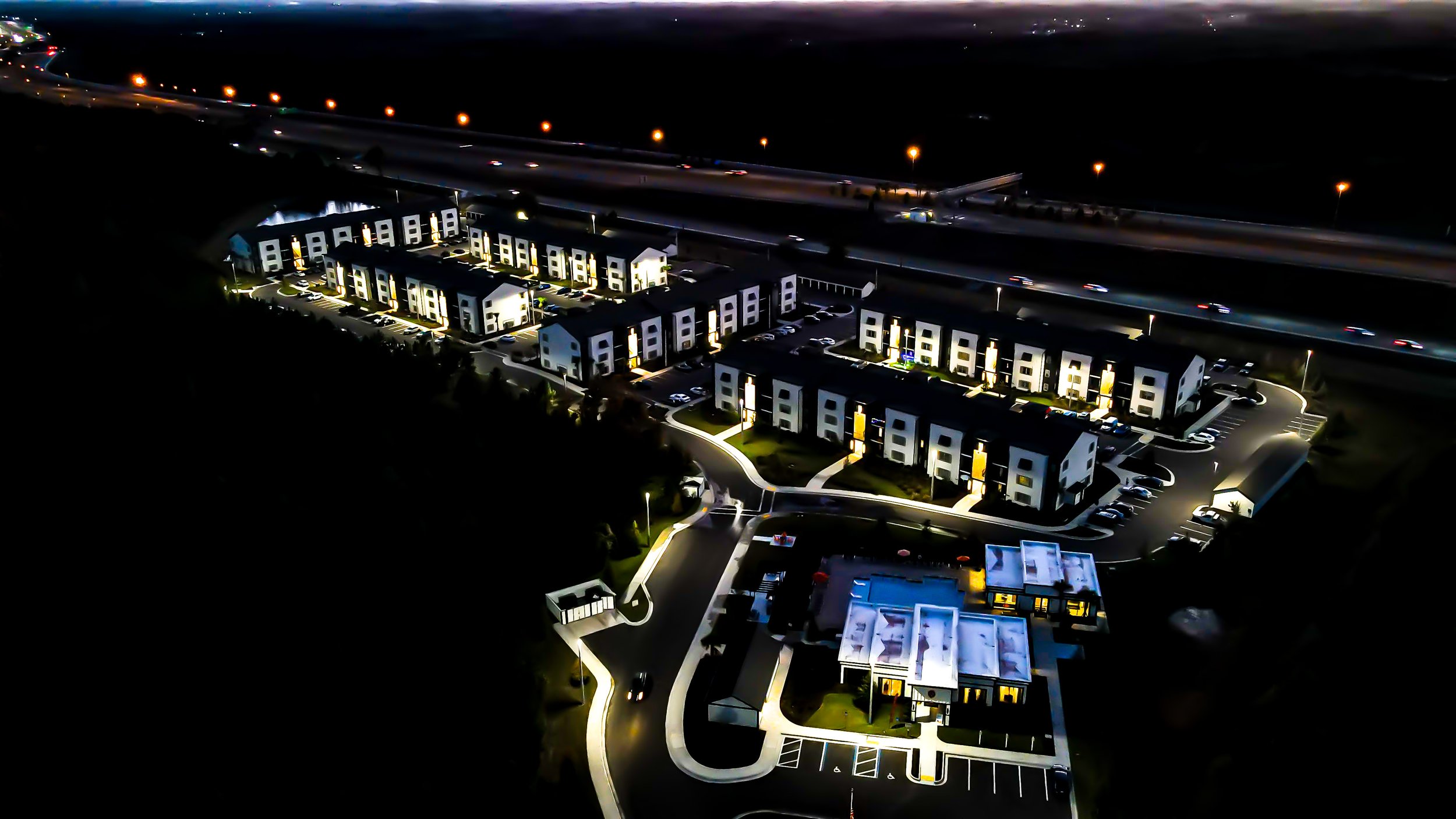 Night aerial view of modern residential apartment complex with illuminated pathways and parking lot, adjacent to a highway with moving cars.