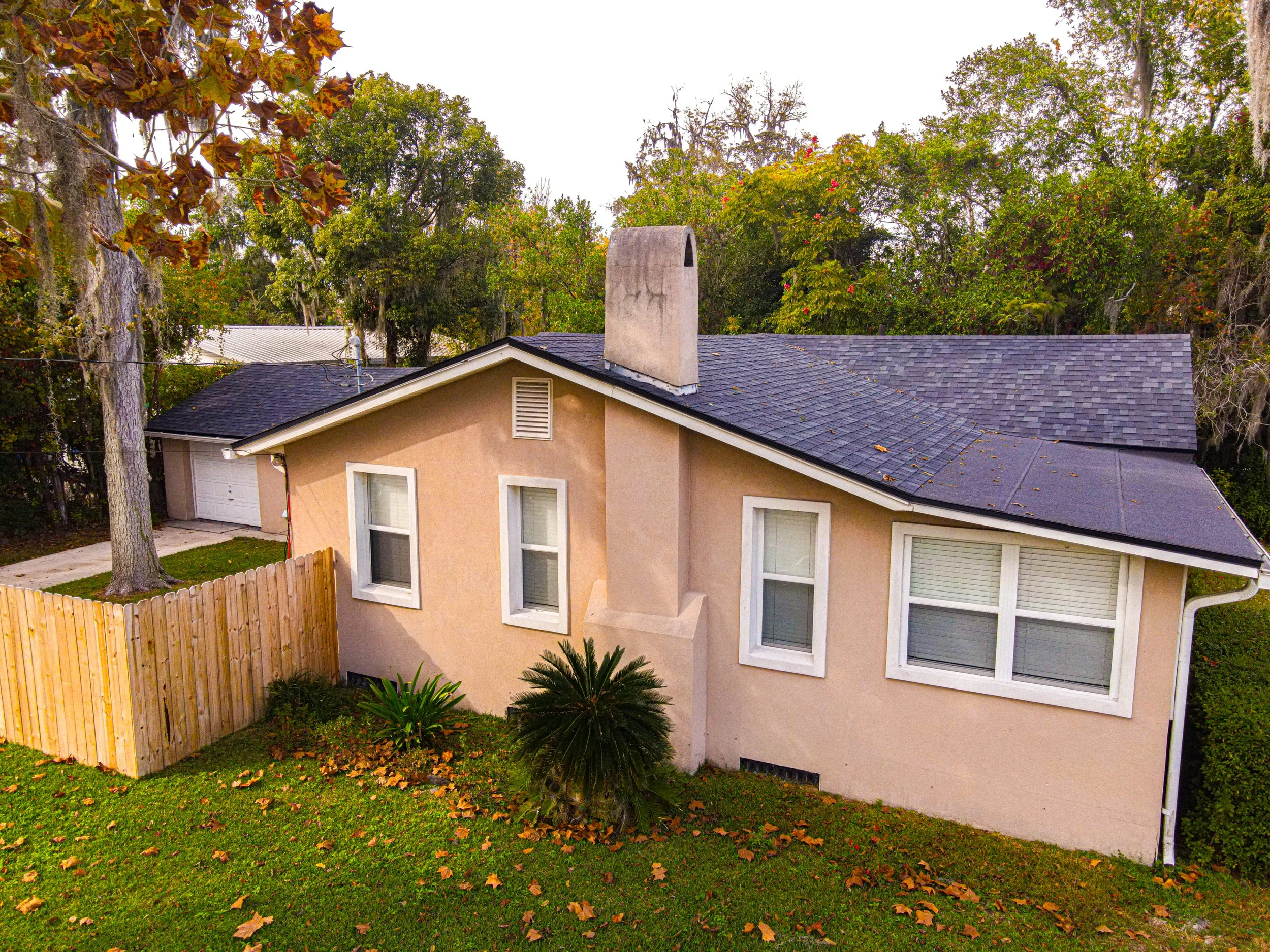 A beige house with a dark blue roof, three windows, and a small fence in the backyard, surrounded by trees with fall foliage.