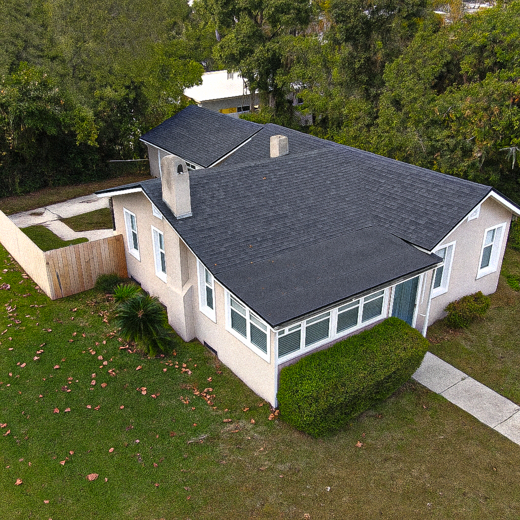 An aerial view of a house with a dark gray roof, white exterior walls, and a front porch area. The house is surrounded by a green lawn with some fallen leaves and is bordered by a wooden fence and tall trees in the background.