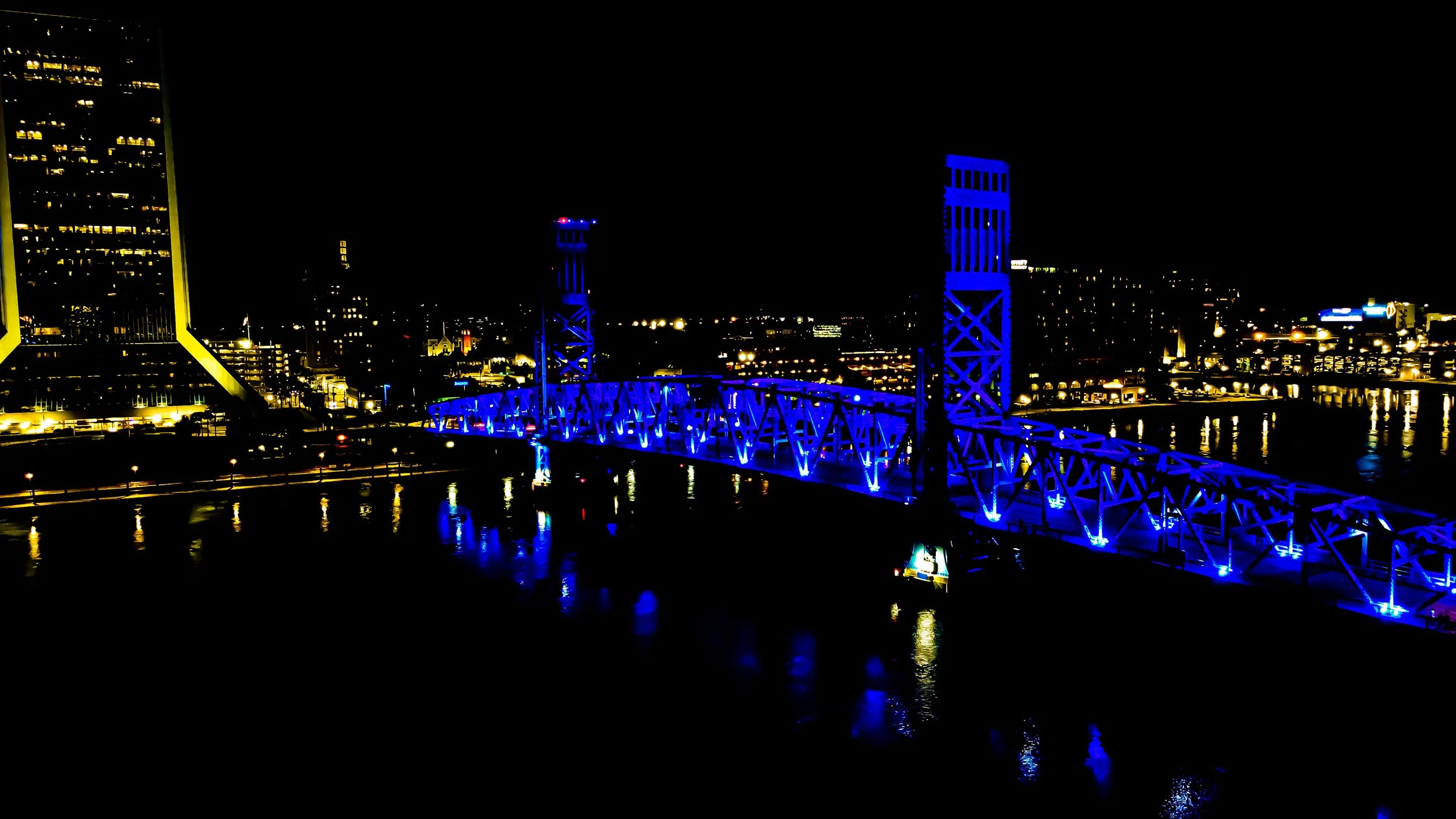 Nighttime cityscape featuring a bridge illuminated with blue lights over a body of water, with buildings lit in the background.