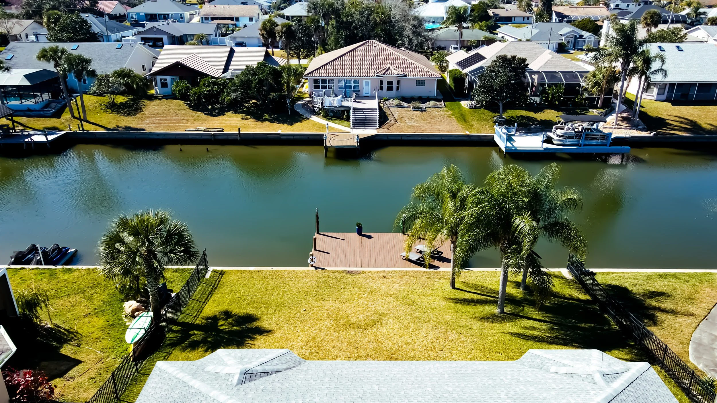 Aerial view of a backyard canal with a dock, palm trees, a grassy lawn, neighboring houses, and boats docked along the water.