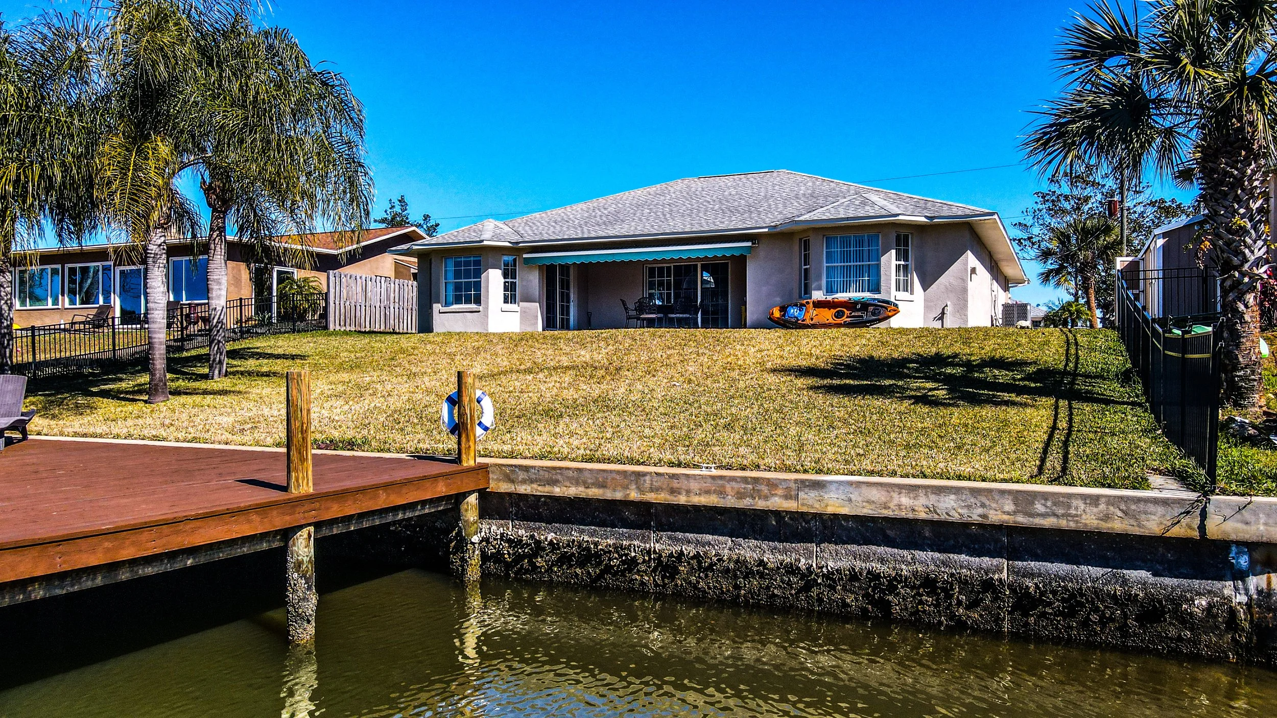 A house with a grassy backyard, palm trees, and a dock by the water under a blue sky.