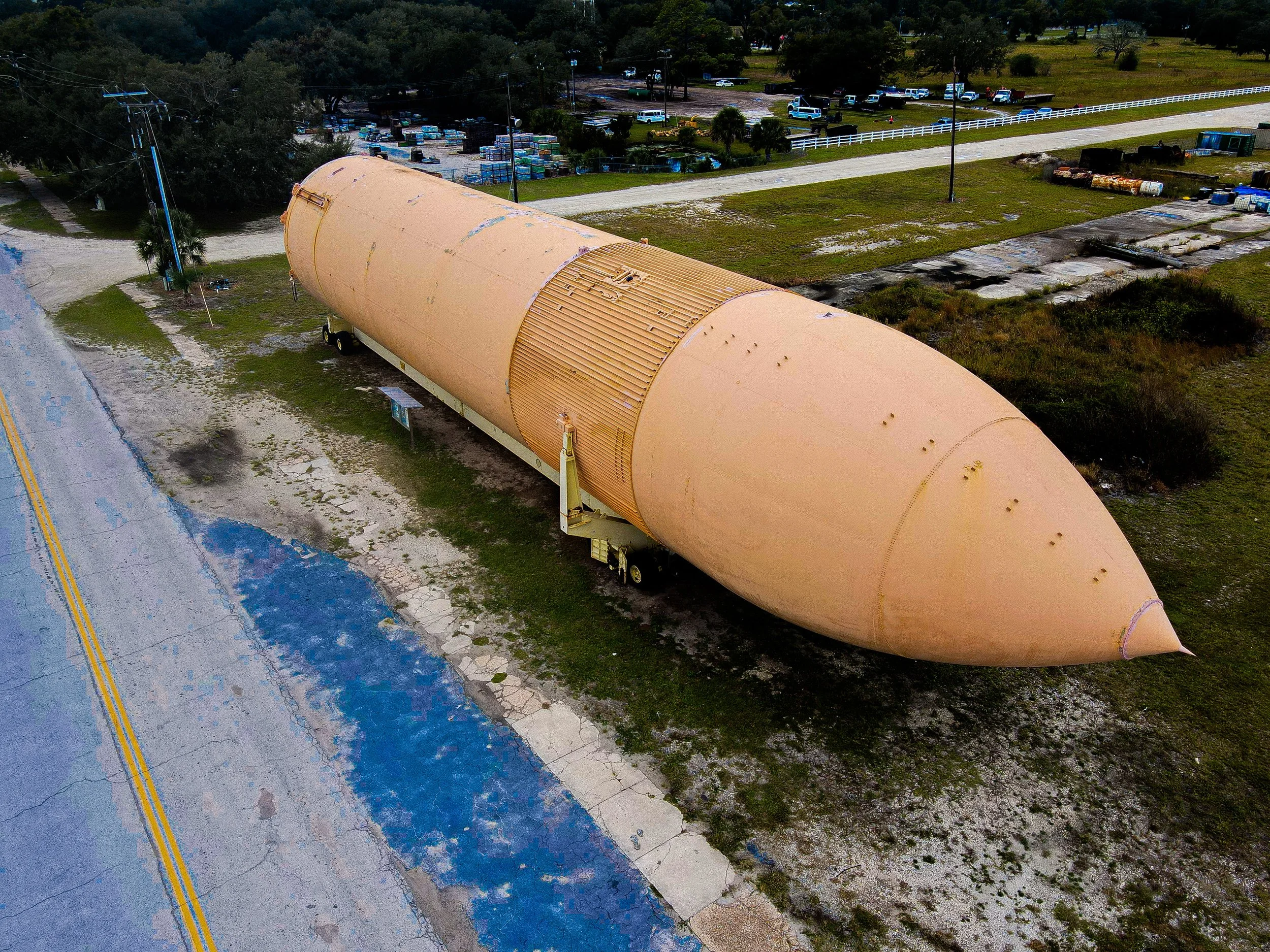 Large orange missile stored outdoors on a truck trailer, parked beside a road with yellow markings. Surrounding area has grass, trees, and miscellaneous items including barrels and vehicles in the distance.