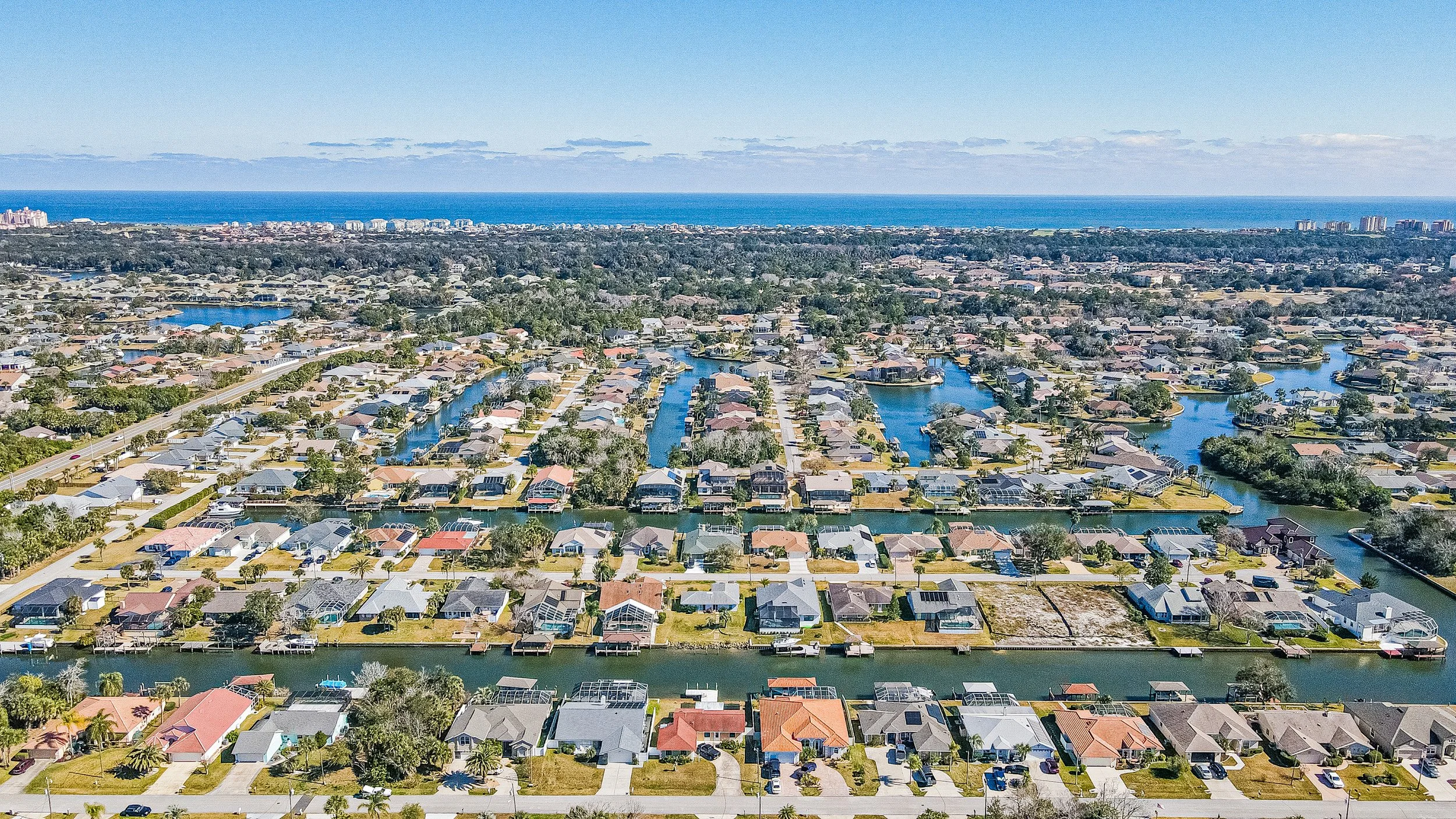 Aerial view of a residential neighborhood with waterways, houses, trees, and a distant ocean shoreline.