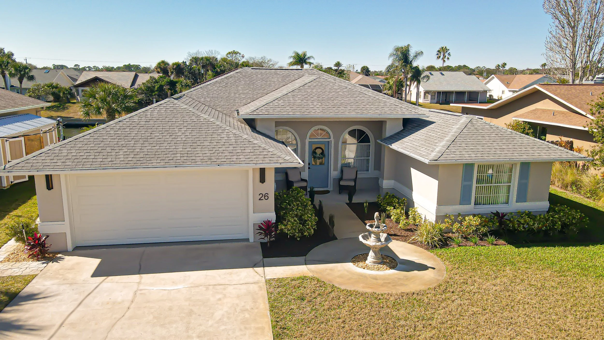 A single-story house with a gray roof and white walls, featuring a two-car garage, a front porch with two chairs, a small fountain, and a well-maintained front yard with plants and a curved walkway.
