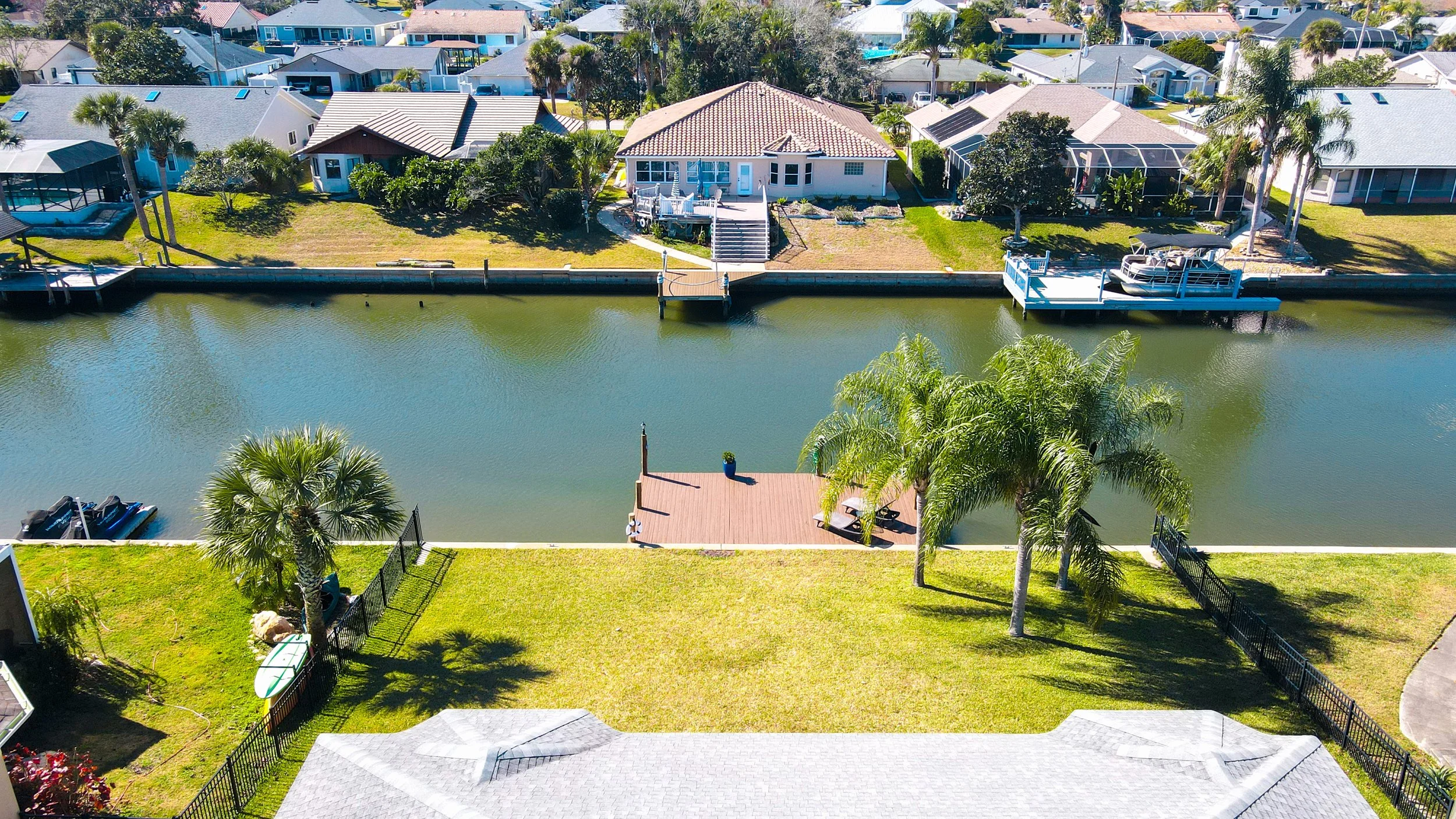 Aerial view of waterfront houses with backyards along a canal, featuring docks, boats, palm trees, and well-maintained lawns.