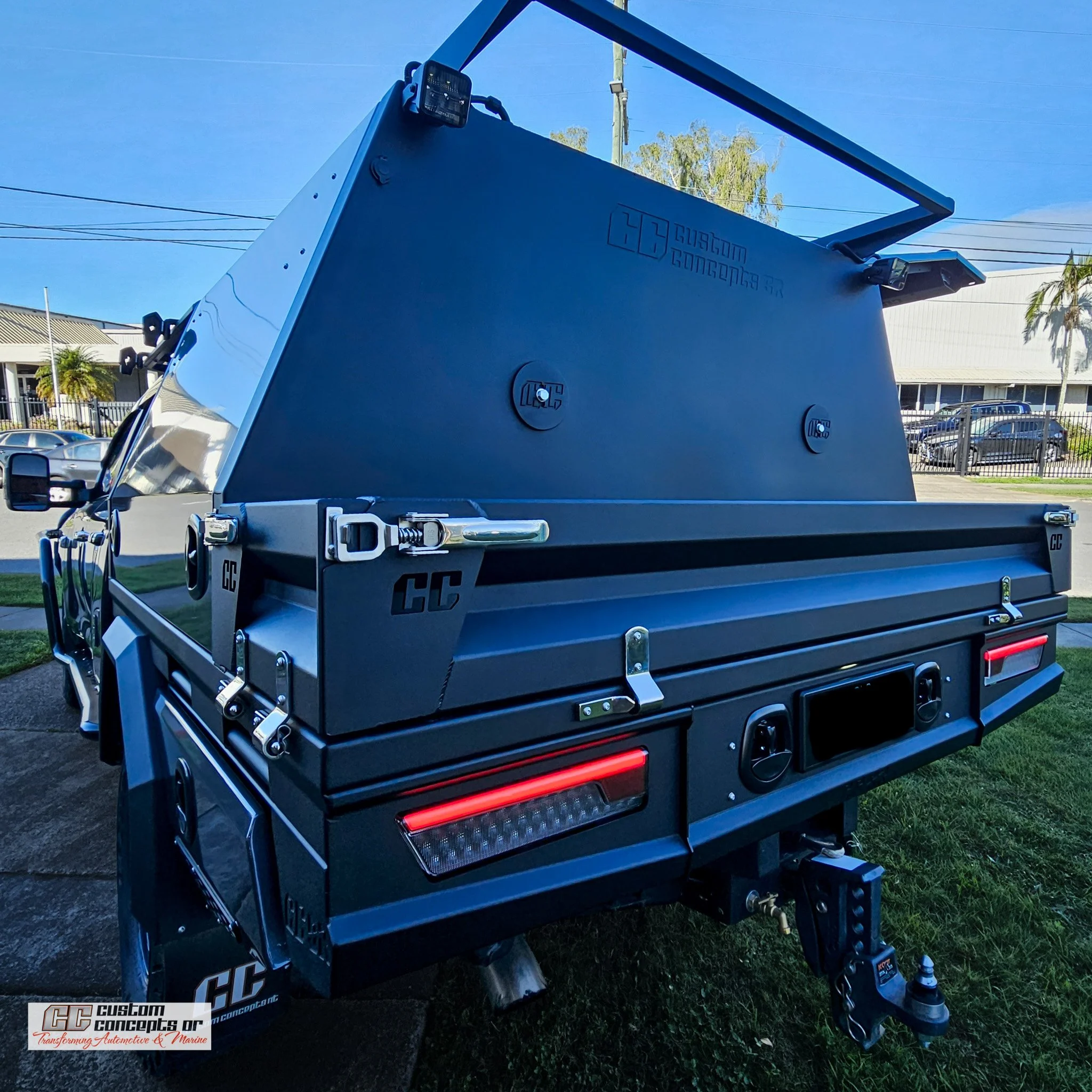 Close-up of a large automotive or marine battery box with metal hardware, mounted on a vehicle, with power connectors and cables, outdoors under a blue sky with buildings and trees in the background.