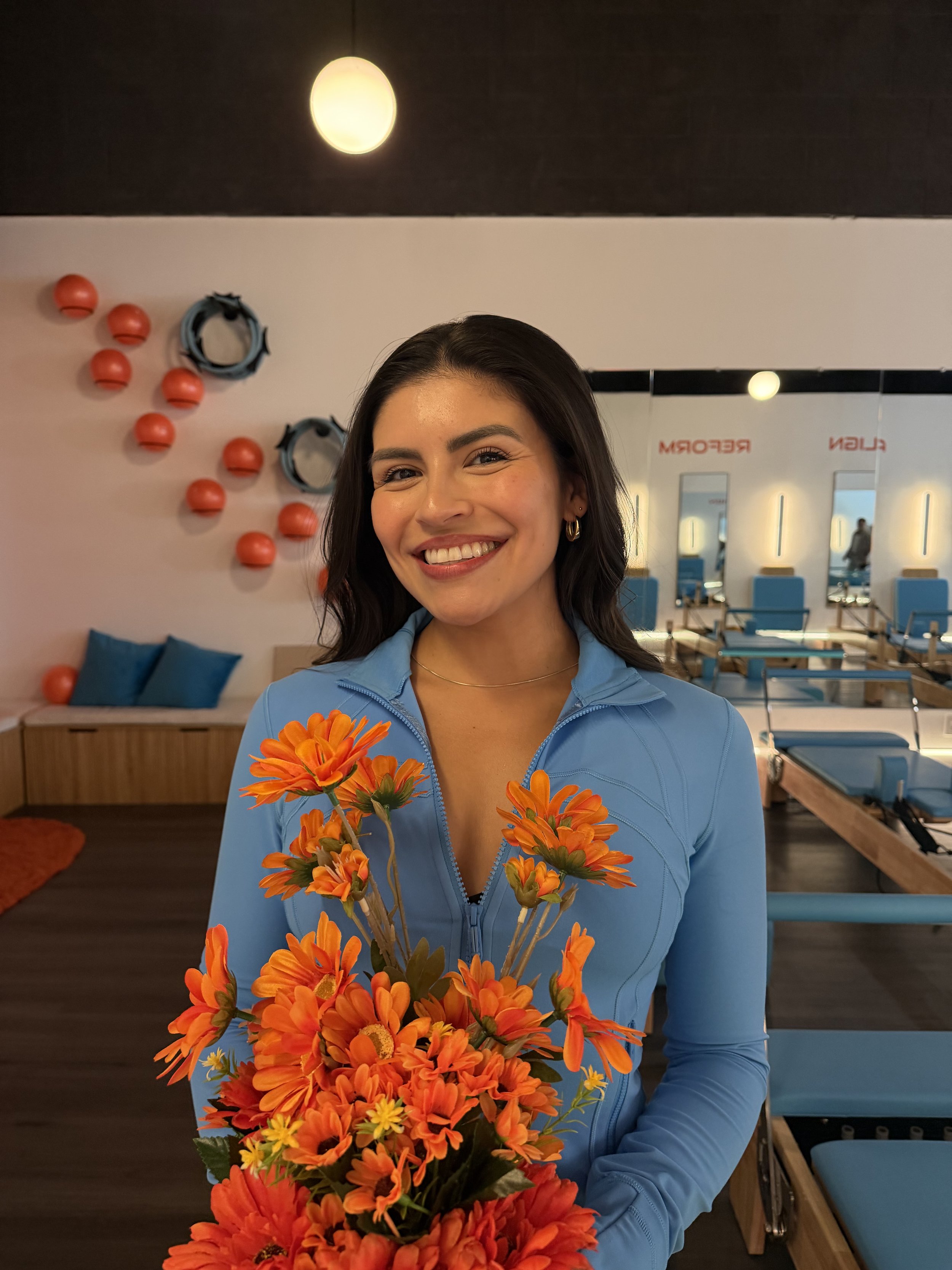 A woman smiling and posing with her hands on her hips, wearing a blue turtleneck shirt, standing against a plain, light-colored background.