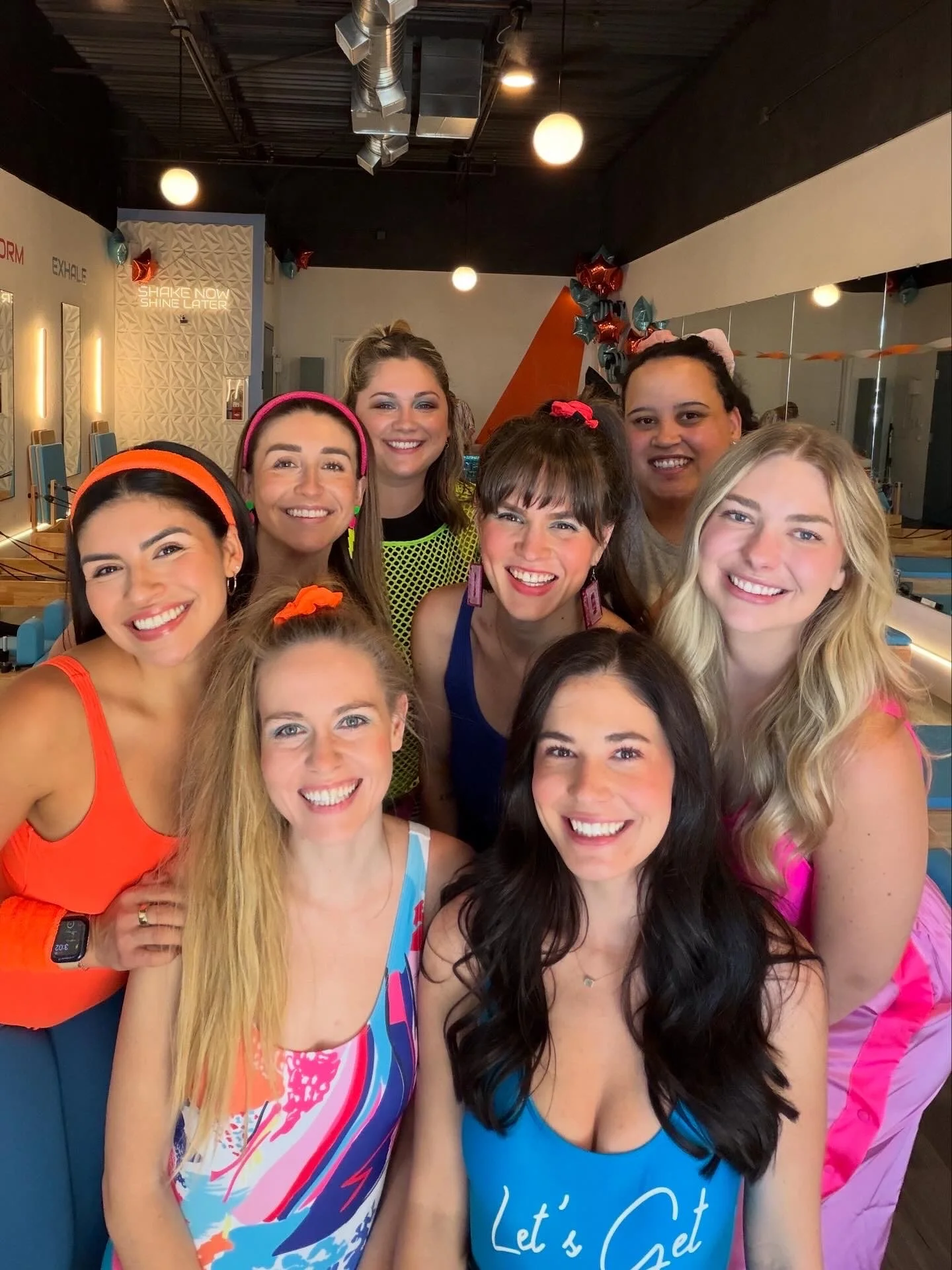 Group of nine young women smiling indoors, wearing colorful outfits and accessories, with a decorated wall and mirror behind them.