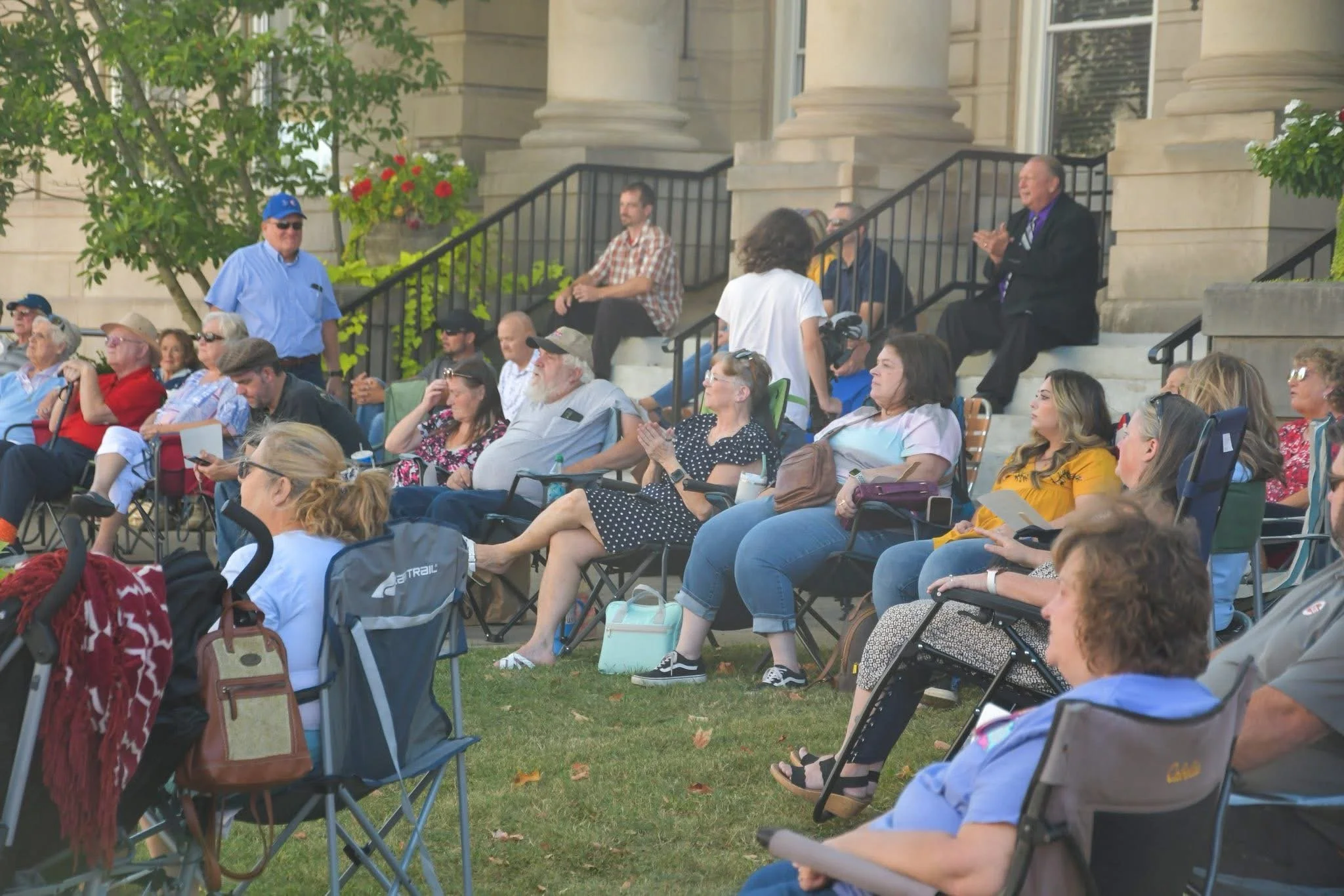 A group of people gathered outdoors, seated on chairs and benches, attending an event in front of a historic building with columns. Some are clapping, others are using devices or talking, with a man in a suit sitting on the stairs.
