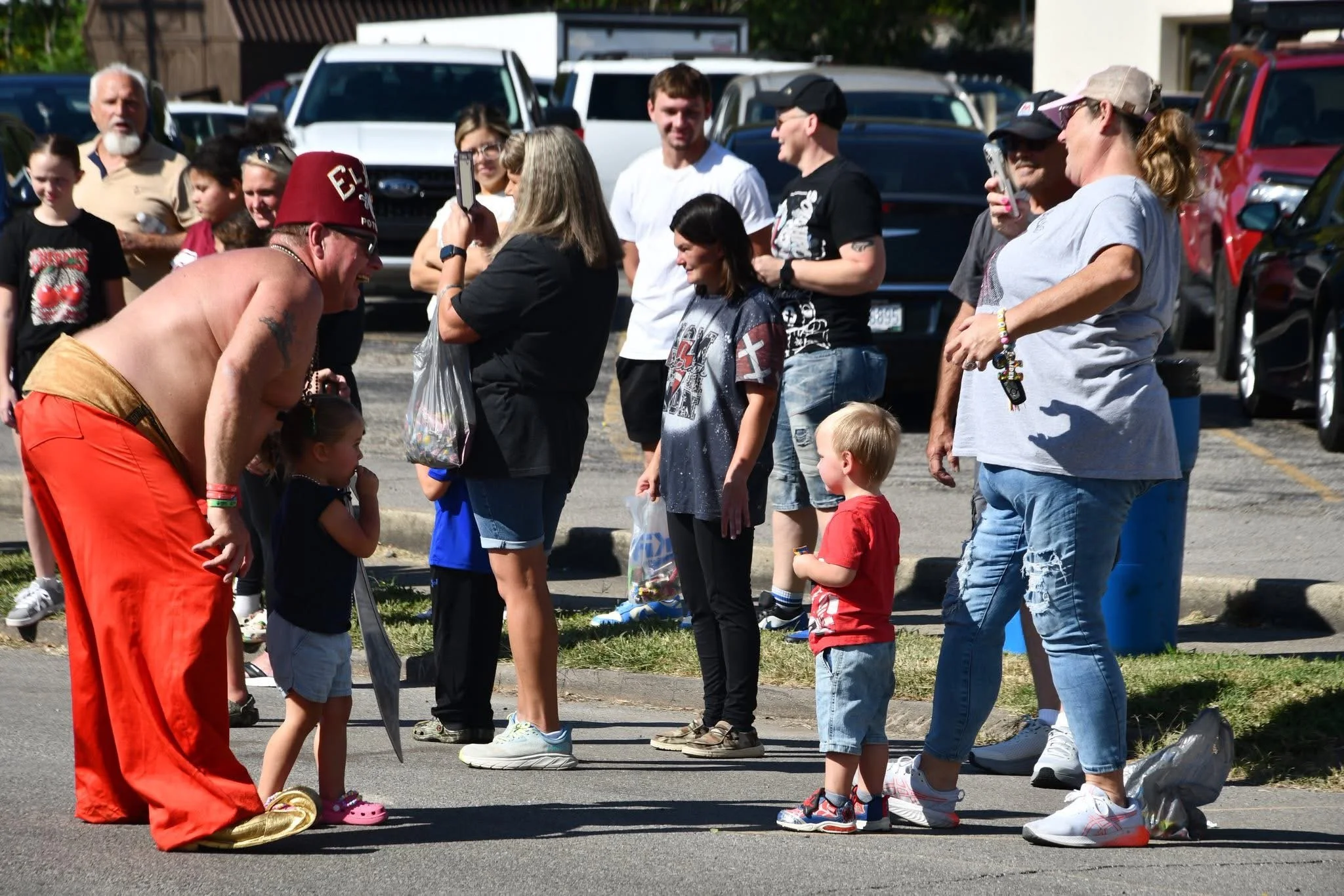 A shirtless man dressed as a clown or performer with a red hat and orange pants bending down to interact with two young children, in a parking lot with a group of onlookers, some taking photos, and parked cars in the background.