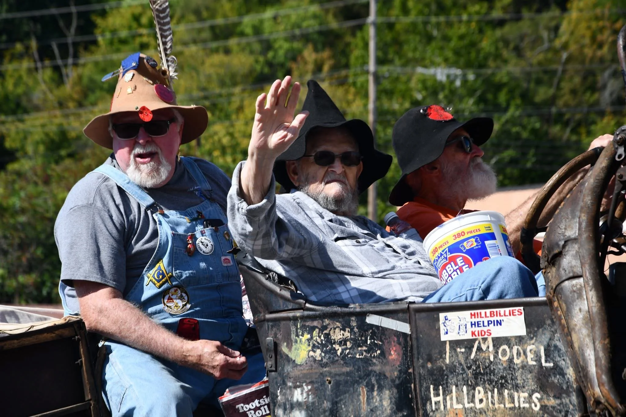 Three older men riding in a rusty vehicle during a parade, wearing black hats and sunglasses. One man is waving and holding a bucket of popcorn, with stickers on the side of the vehicle, including one that says "HILLBILLIES HELPIN' KIDS".