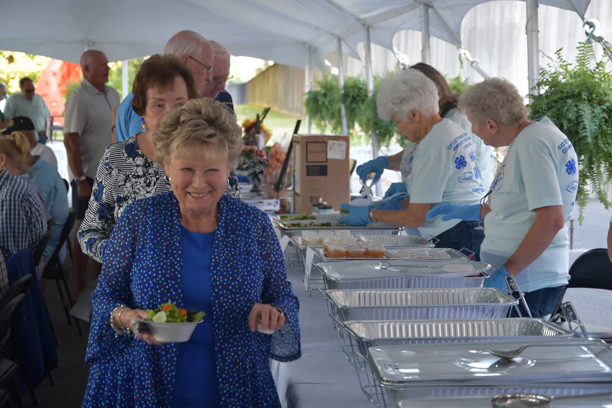 A woman smiling and holding a bowl of salad in front of a buffet line at an outdoor event under a white tent, with elderly women serving food and other people in the background.