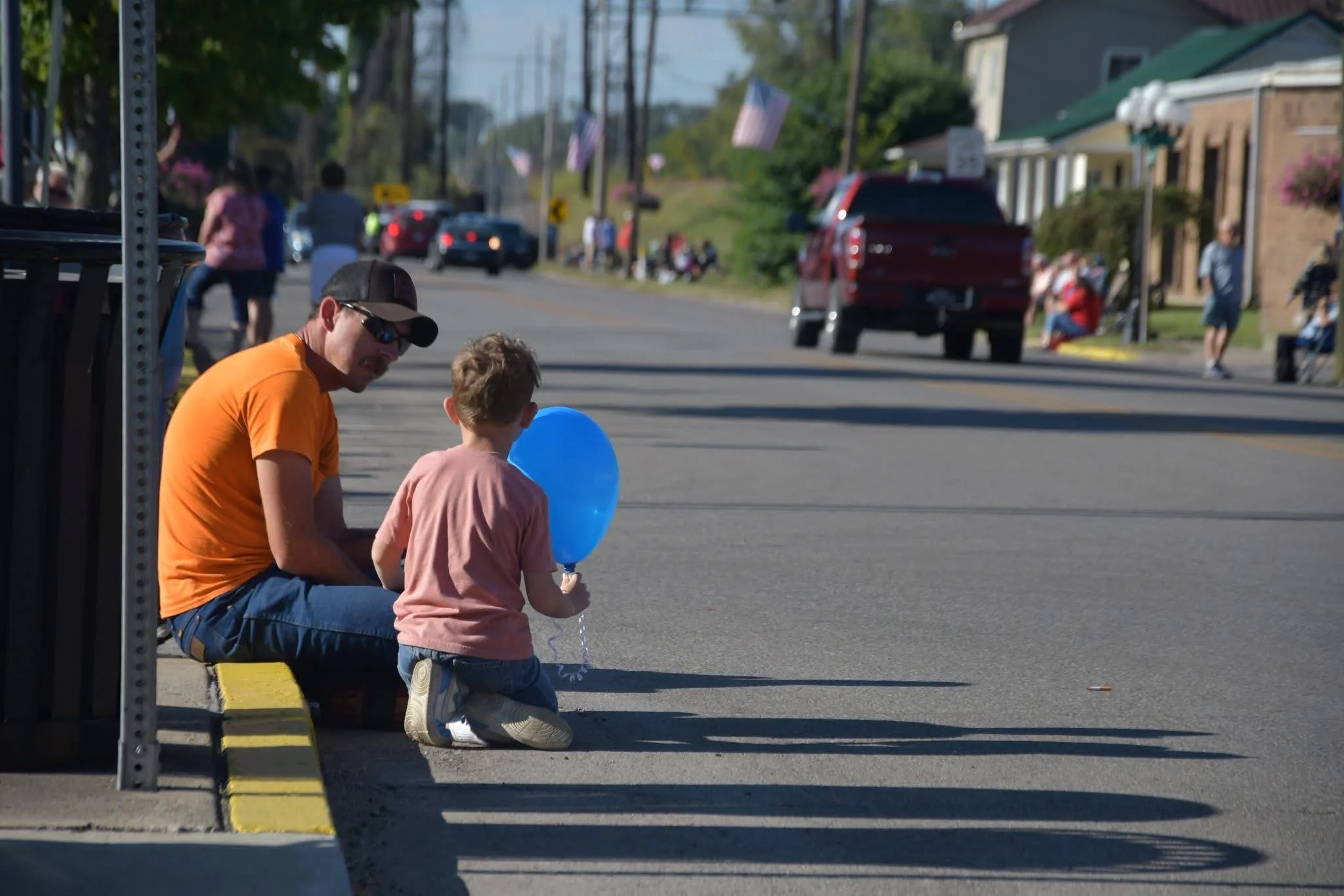 A man and a young boy sit on the sidewalk at a street corner, with the boy holding a blue balloon. They are shaded by a street sign and are surrounded by an active street scene with people, cars, and buildings in the background.