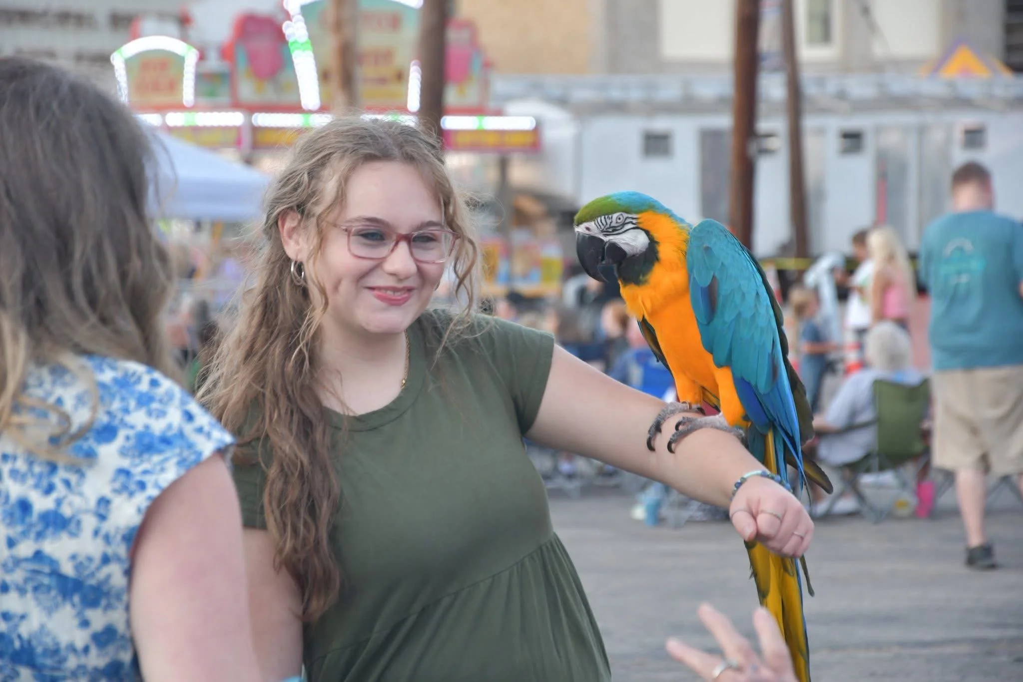A woman with glasses and long wavy hair smiling, with a colorful blue and yellow macaw perched on her arm, at an outdoor fair or festival with bright lights and people in the background.