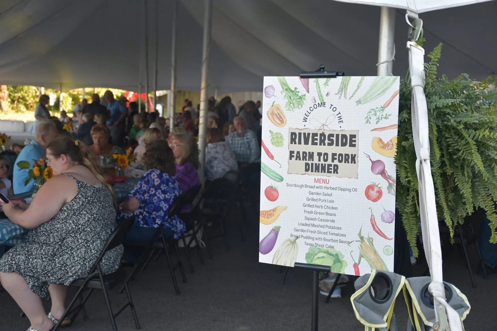 People dining under a large tent at a farm-to-fork dinner event, with a menu sign featuring vegetables and dishes such as garden salad and grilled pork loin in the foreground.