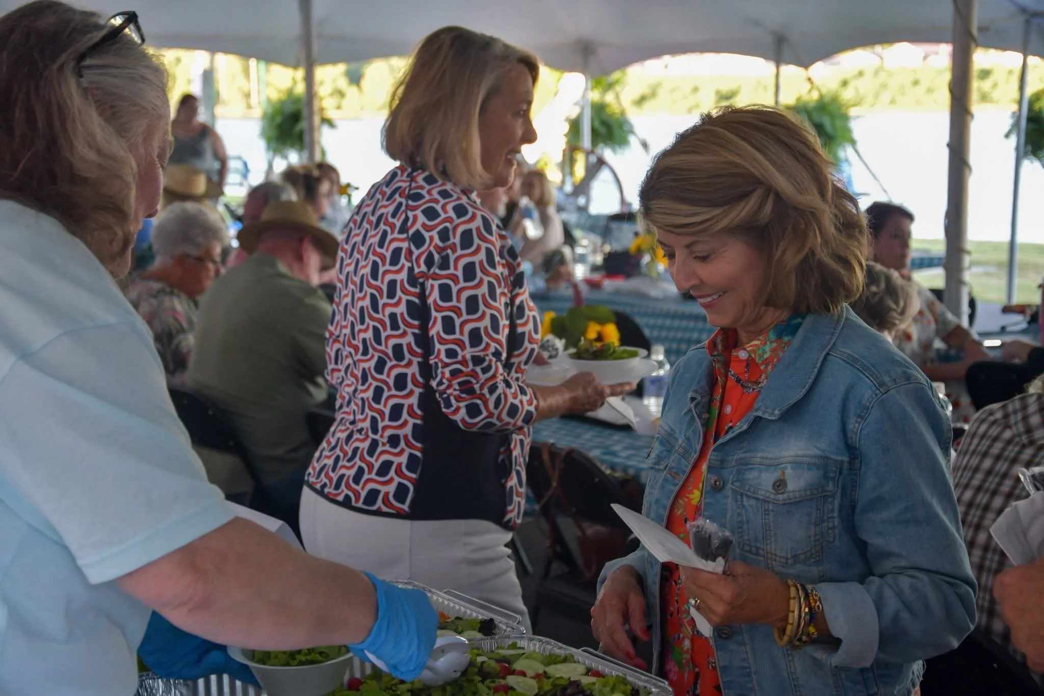 People at an outdoor gathering, serving themselves food from a buffet table under a tent, with greenery and water in the background