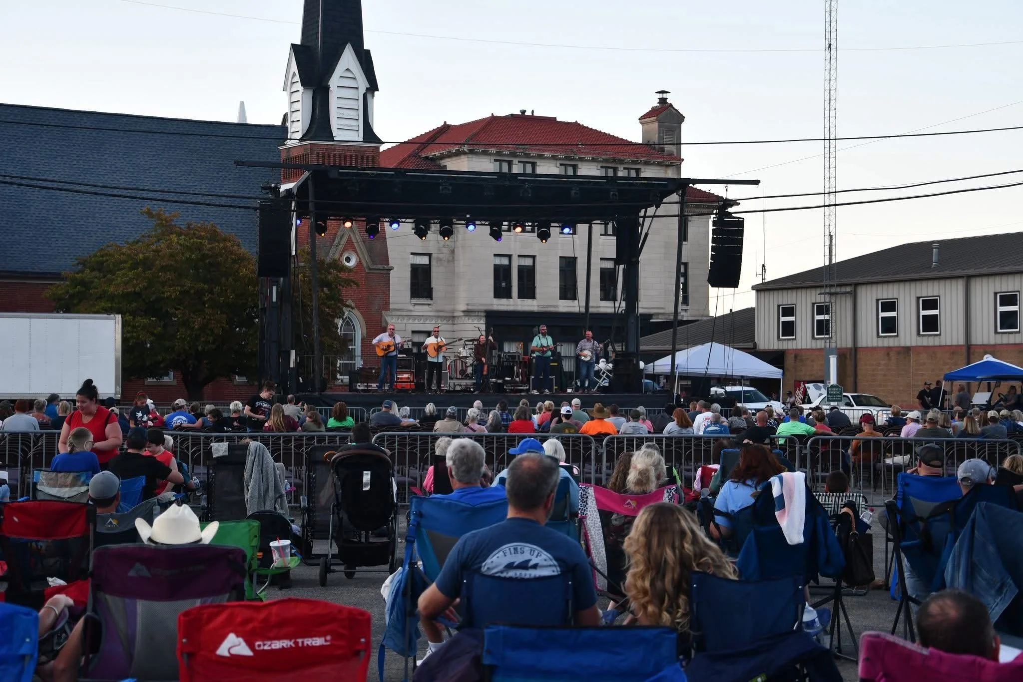 An outdoor concert in front of a historic building with a red roof. The stage has musicians playing guitars, and the audience is seated in chairs and blankets, watching the performance.