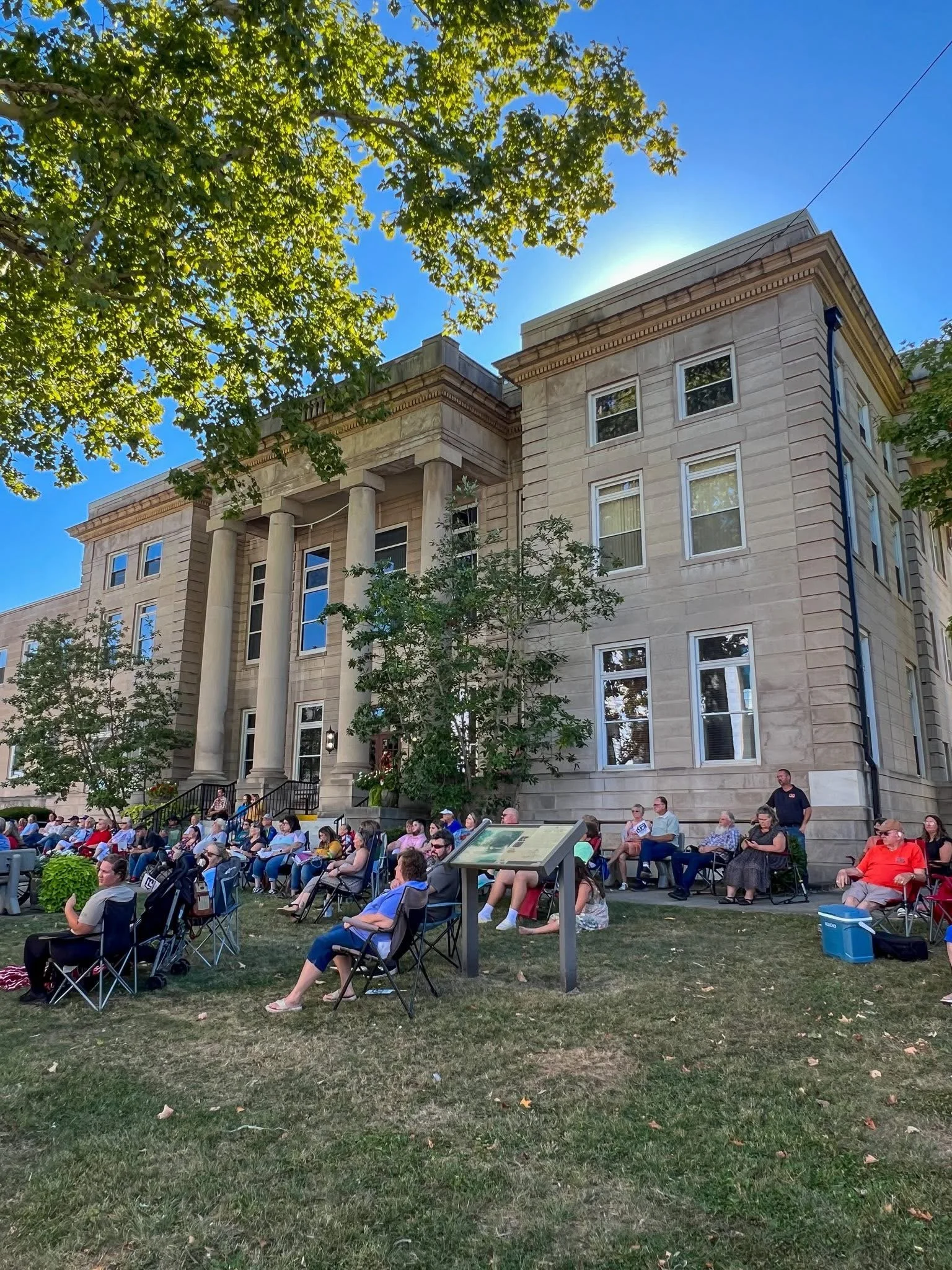 People sitting on chairs outdoors in front of a historic stone building with large columns, under a tree on a sunny day.