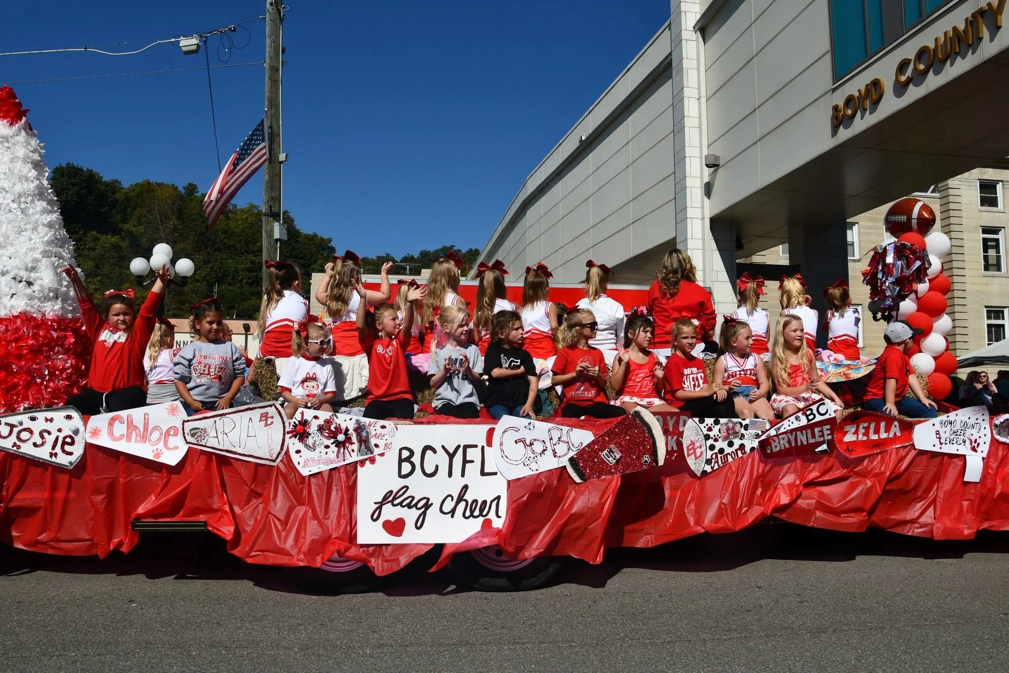 A parade float decorated with red, white, and black items featuring a group of young girls dressed in red and white, some sitting and some standing, participating in a parade. The float includes signs with names and phrases, balloons, and a football 