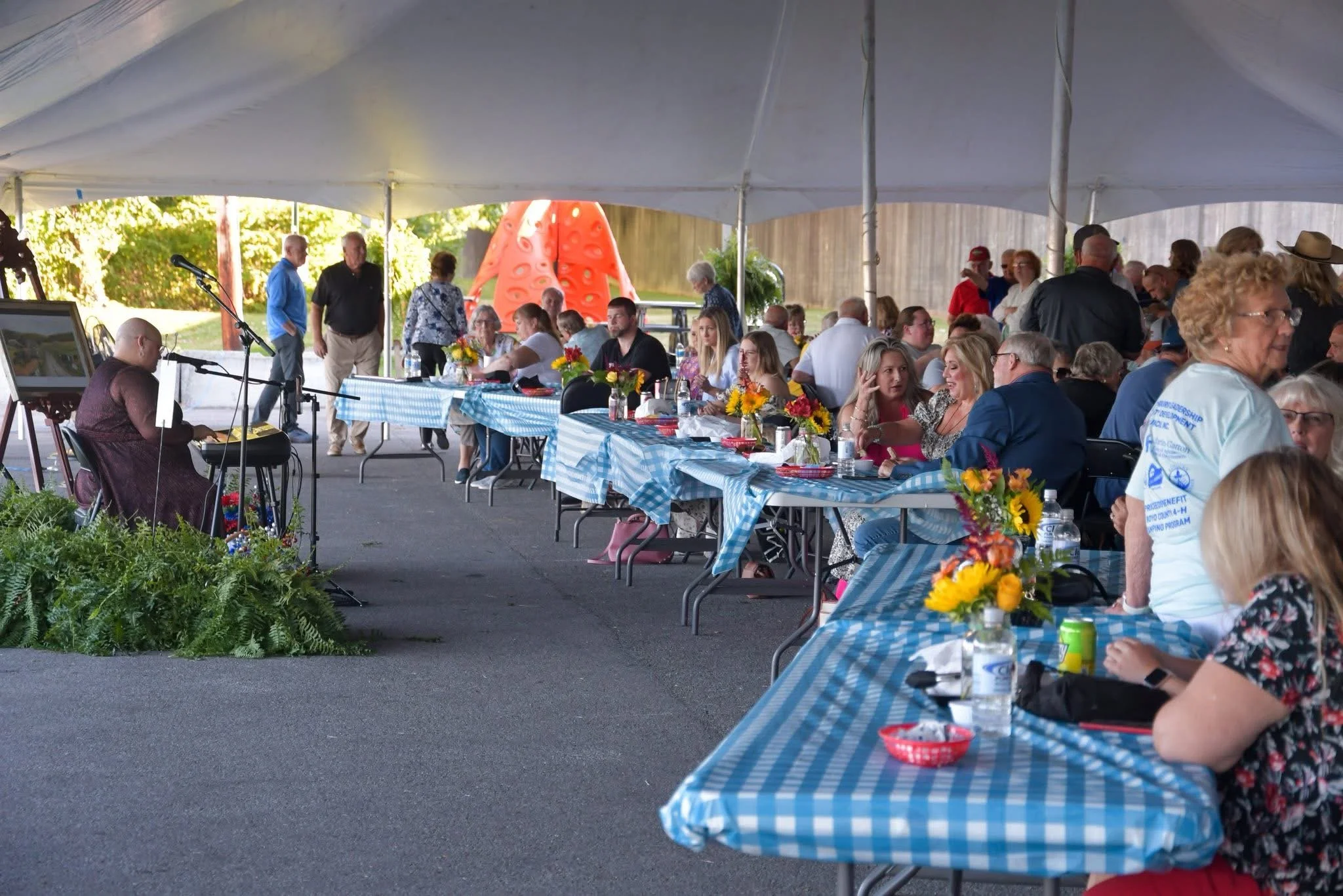 People gathered under a large tent for an outdoor event, sitting at long tables with blue-and-white checkered tablecloths, decorated with yellow and orange flower centerpieces, and watching a performer playing a keyboard near some plants.